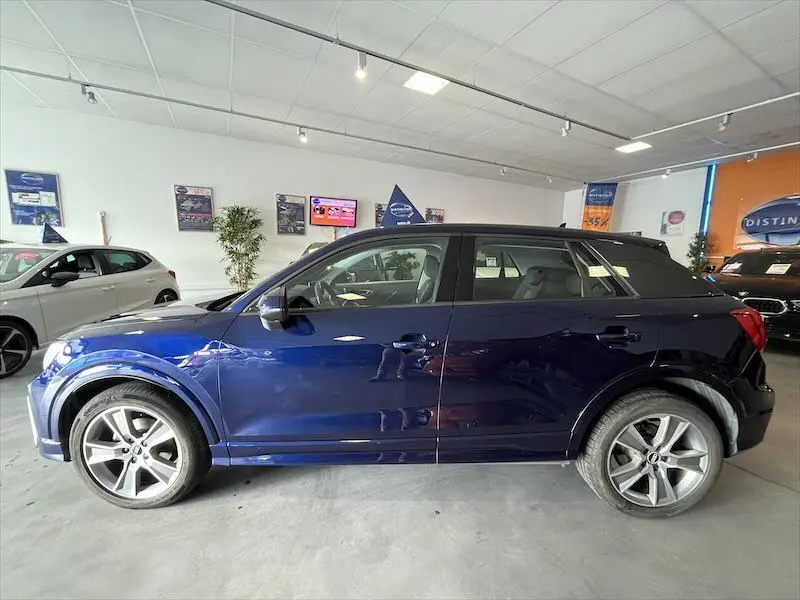 Profil côté gauche d'un Audi Q2 S-Line 2021 en bleu Navarre métal, avec jantes alliage et toit noir en showroom.