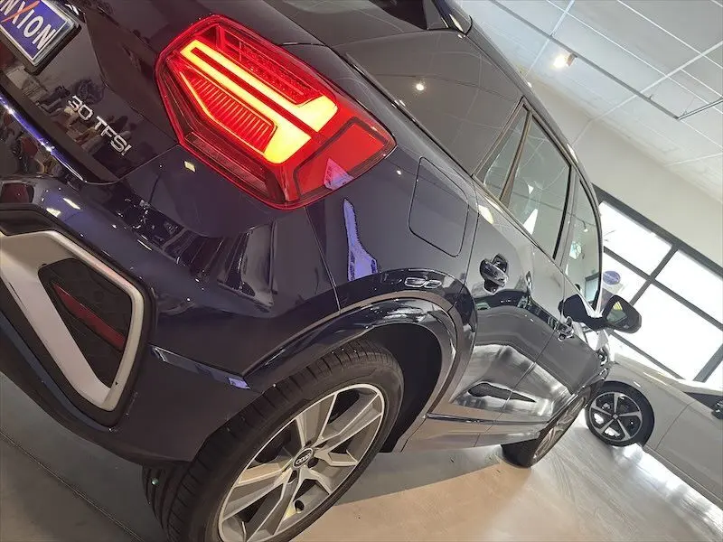Vue 3/4 arrière droite d'un Audi Q2 30 TFSI S-Line bleu Navarre métal avec feux arrière allumés en showroom.