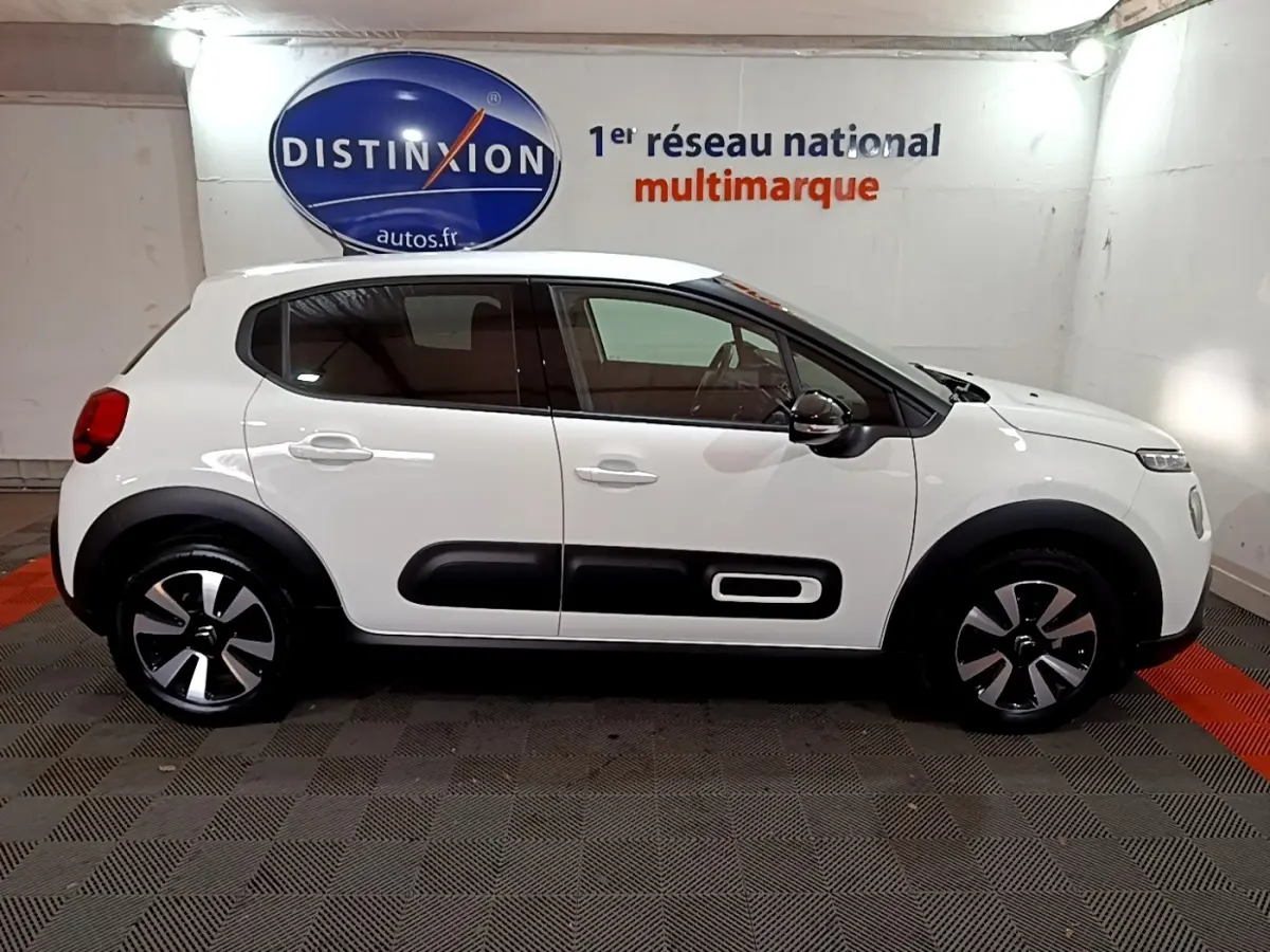 Profil côté droit d'une Citroën C3 blanche 2023 avec protections noires et jantes bicolores en intérieur showroom.