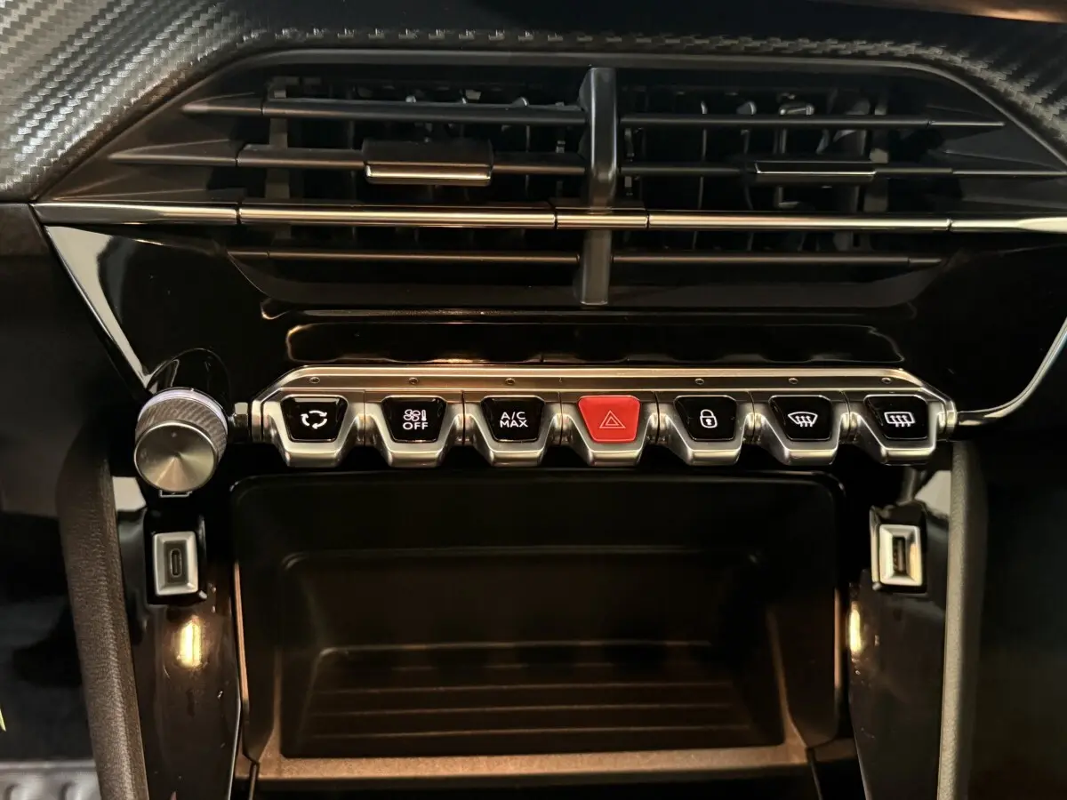 Vue rapprochée du tableau de bord noir brillant du Peugeot 2008 avec rangée de boutons métalliques et bouton rouge d’alerte.