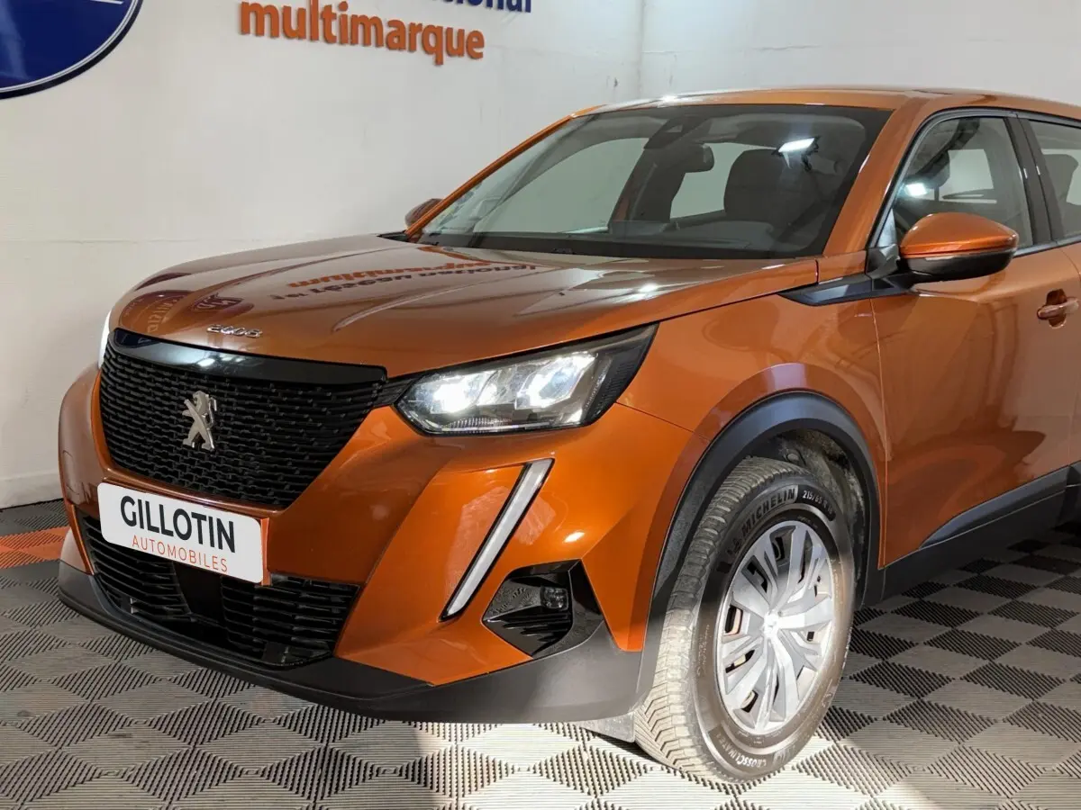 Vue 3/4 avant droit d’un Peugeot 2008 orange 2022 avec calandre noire et feux LED allumés en intérieur showroom.