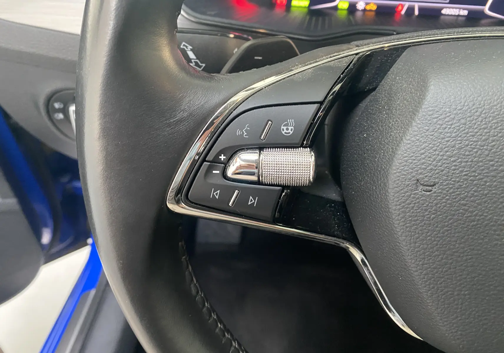 Gros plan sur les commandes gauche du volant en cuir noir du Skoda Superb Combi bleu énergie, avec molette chromée et tableau de bord numérique.