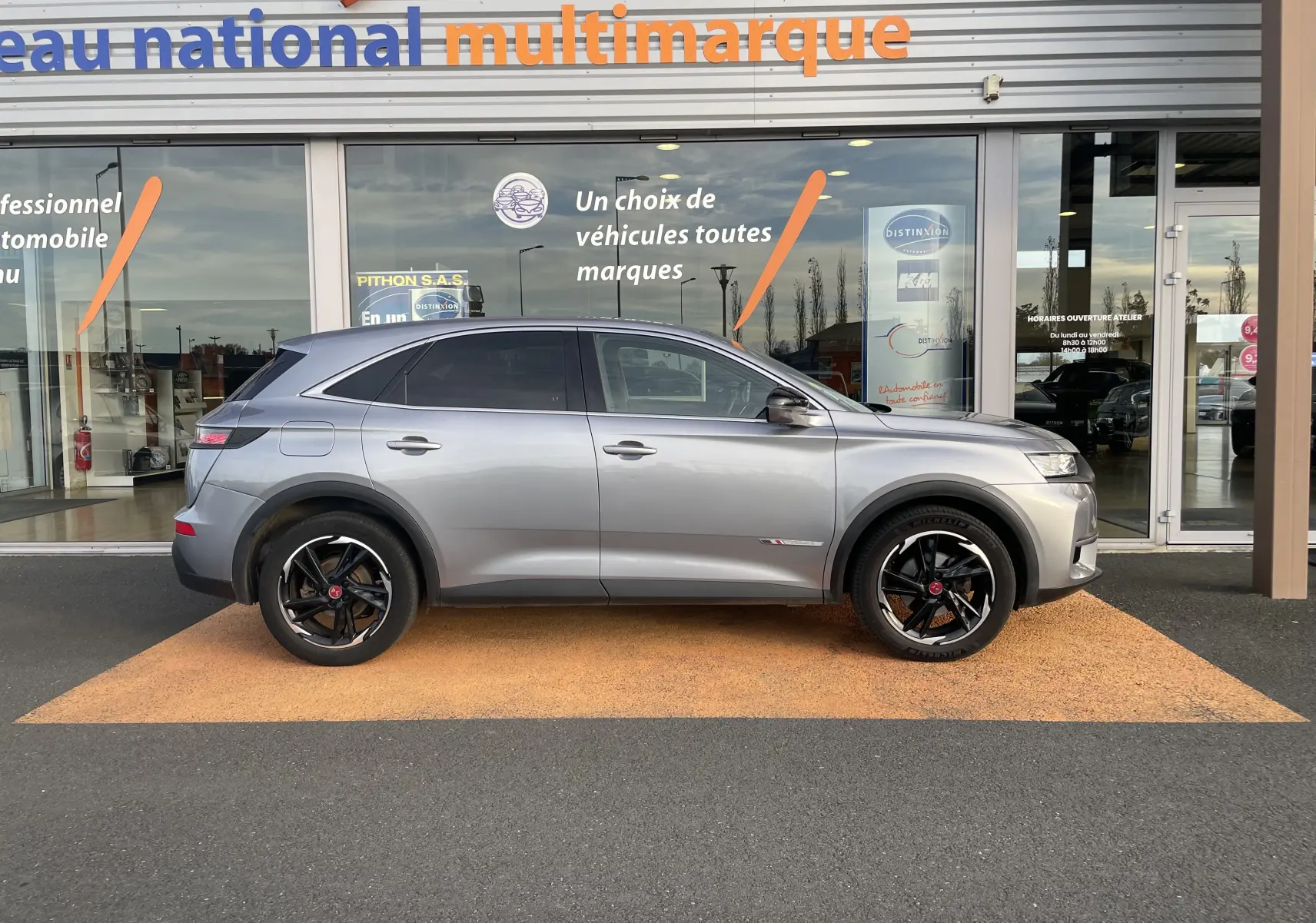 Profil côté gauche d'un DS7 gris 2021 avec jantes noires bicolores, garé devant une vitrine multimarque.