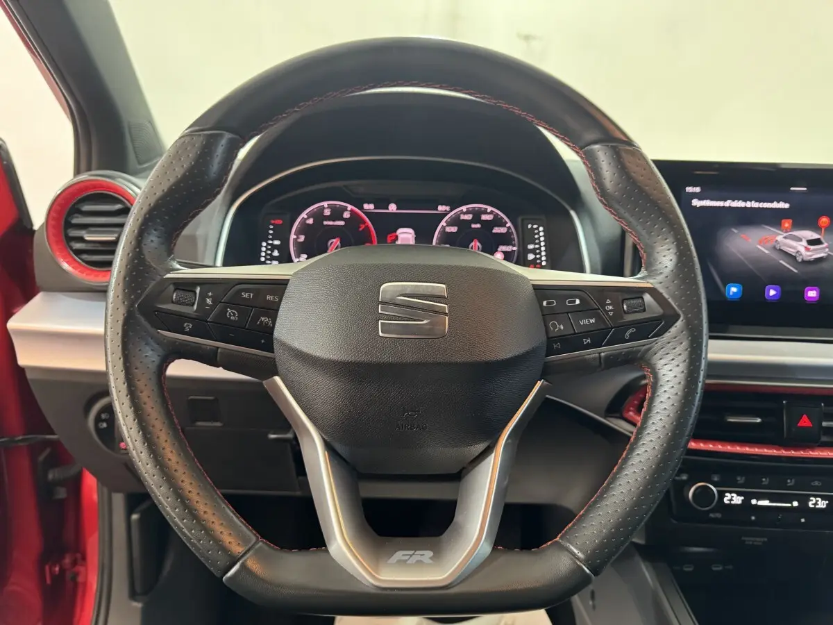 Vue rapprochée du volant cuir perforé avec logo SEAT et badge FR, tableau de bord et écran tactile visibles en arrière-plan.