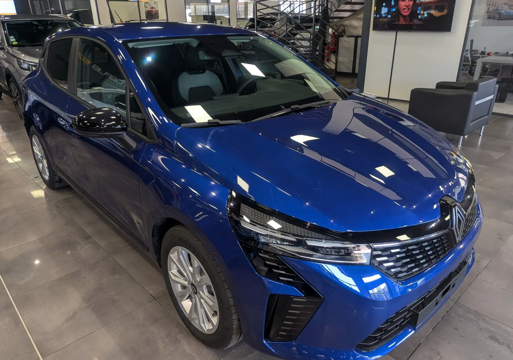 Renault Clio V Bleu Iron vue 3/4 avant droit en intérieur, avec calandre noire et phares LED distinctifs.