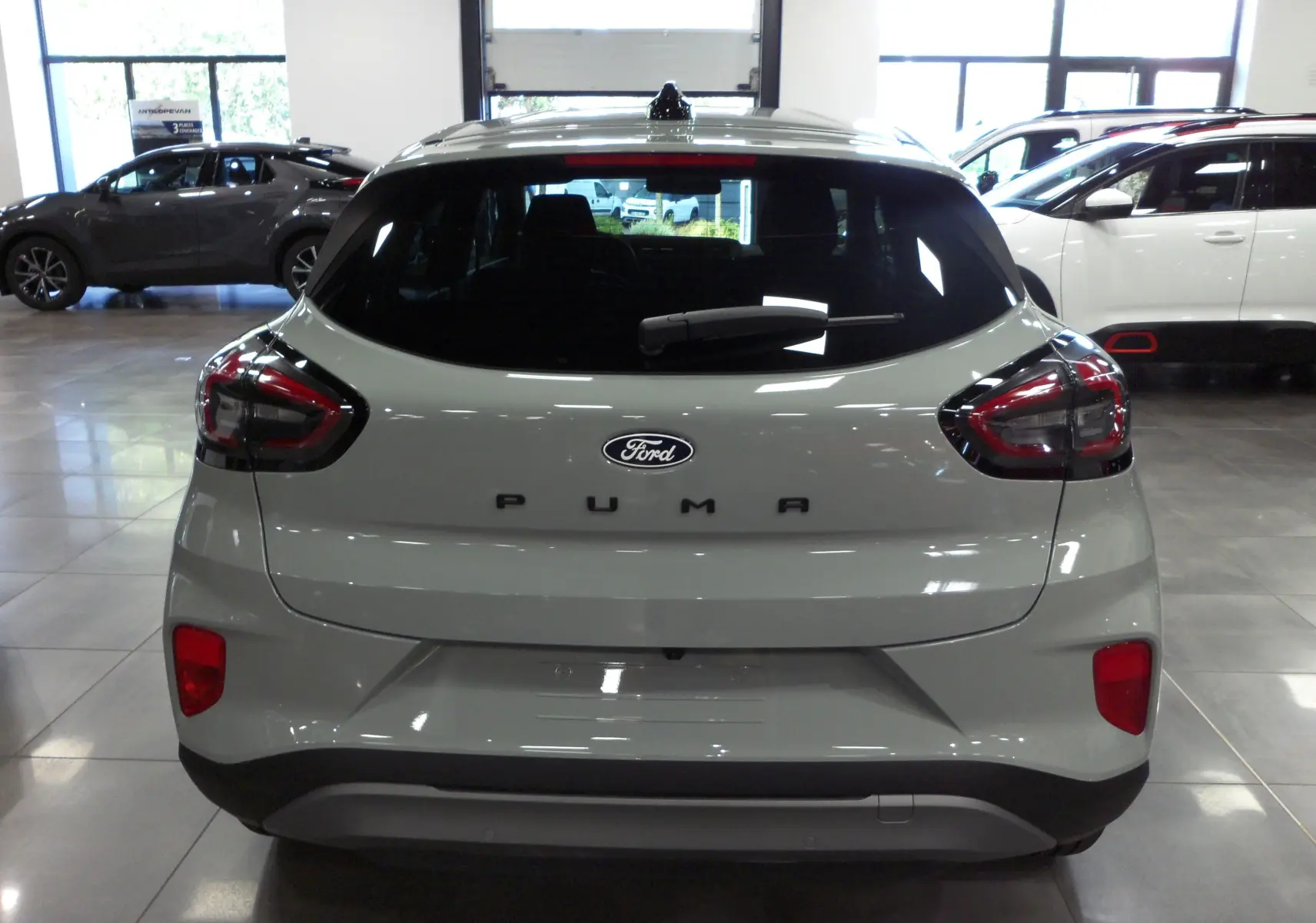 Vue arrière d'un Ford Puma 2025 gris cactus en showroom, avec feux arrière sombres et logo Ford bien visible.