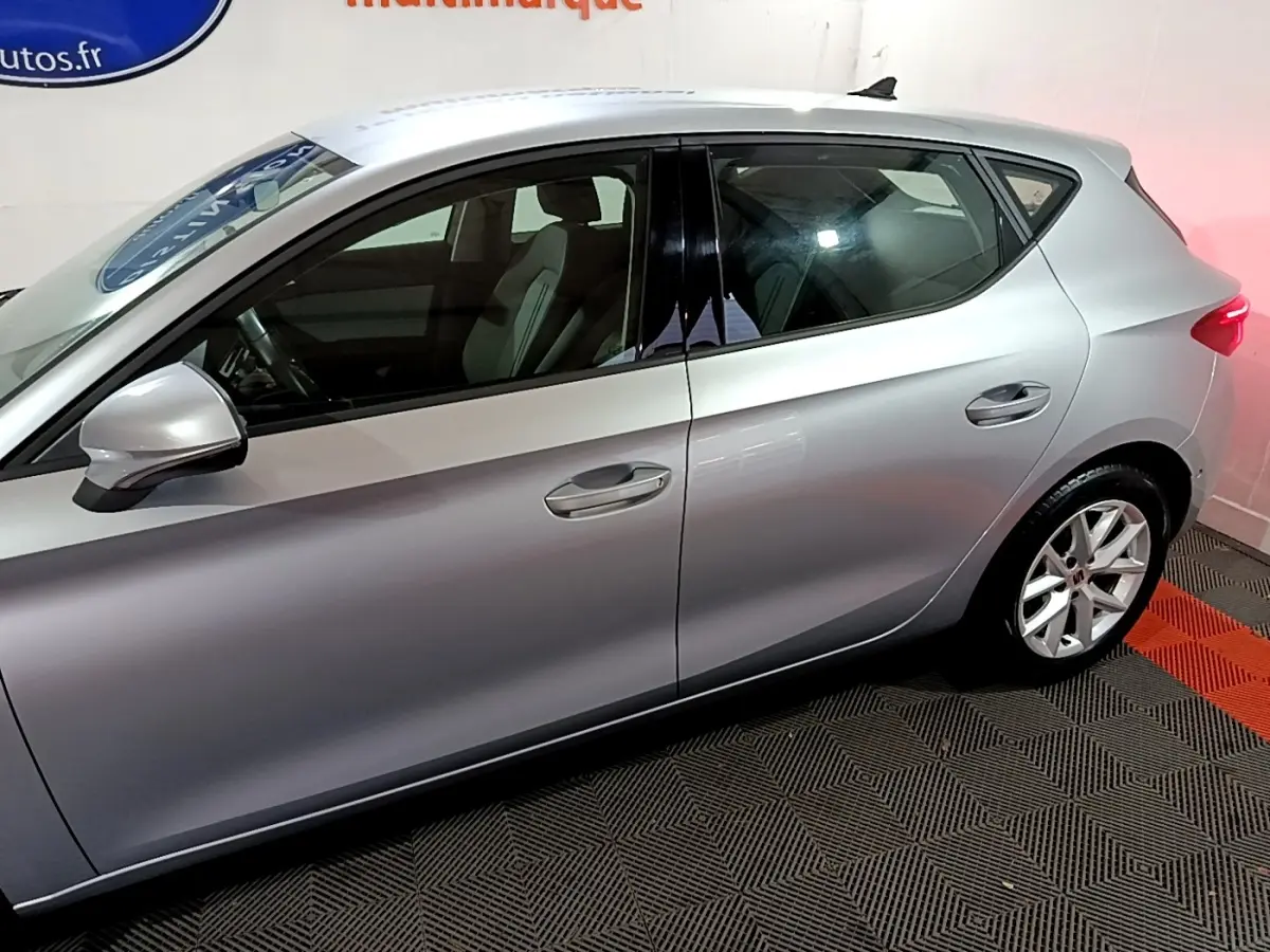 Profil côté gauche d'une SEAT LEON gris clair 2023, avec jantes alliage et vitres teintées dans un showroom.