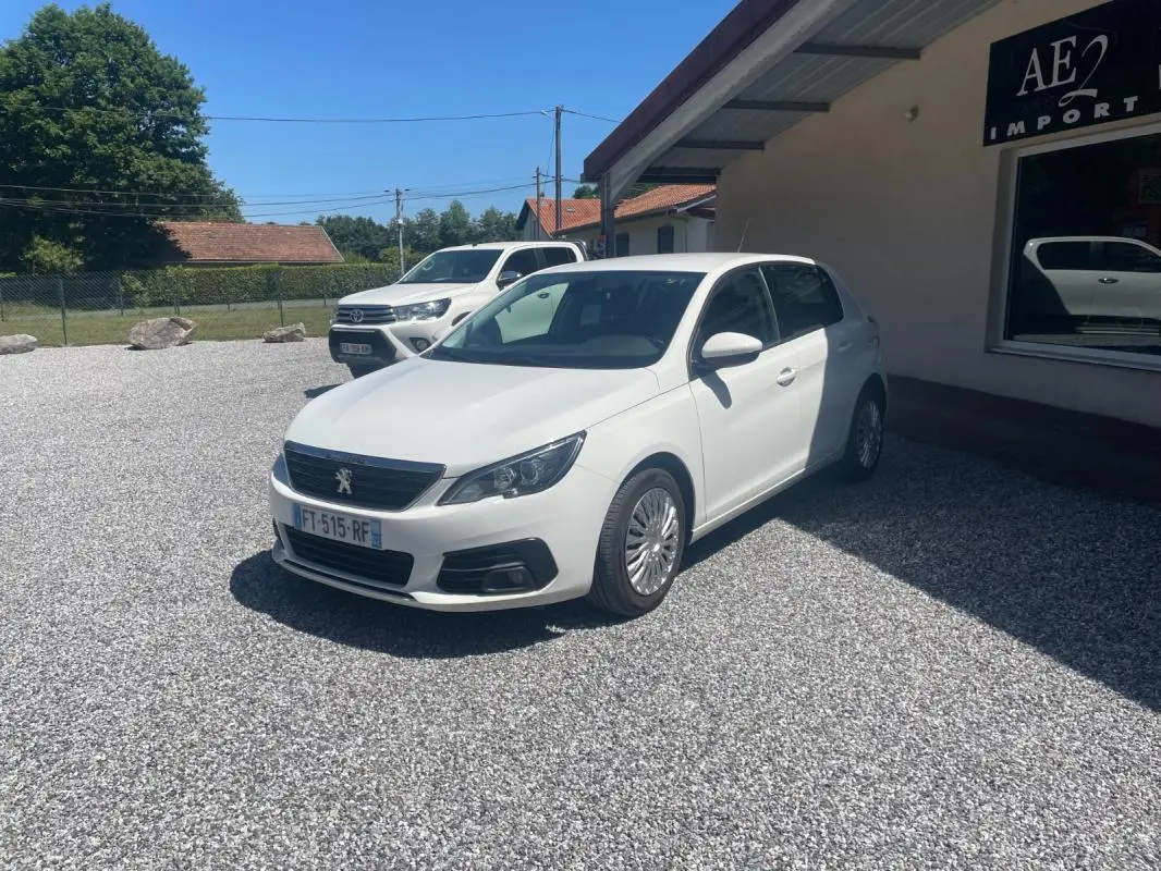 Peugeot 308 blanc vue 3/4 avant droit, garée sur un sol gravillonné devant un bâtiment avec enseigne AE2 Import.