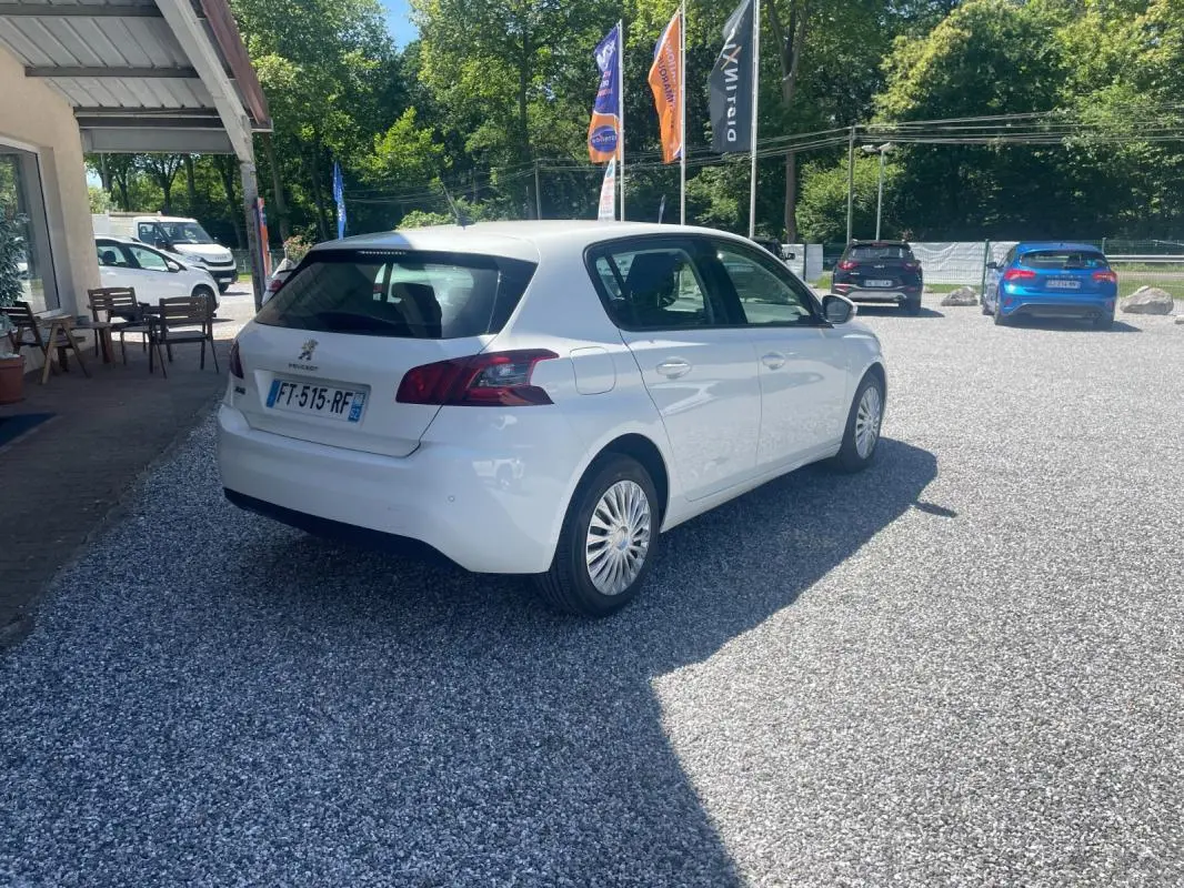 Peugeot 308 blanc vue 3/4 arrière droit sur parking gravillonné avec drapeaux publicitaires en arrière-plan.
