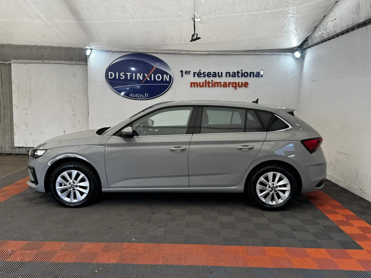 Vue de profil côté gauche d'une Skoda Scala gris clair 2025 dans un showroom avec sol en damier noir et orange.