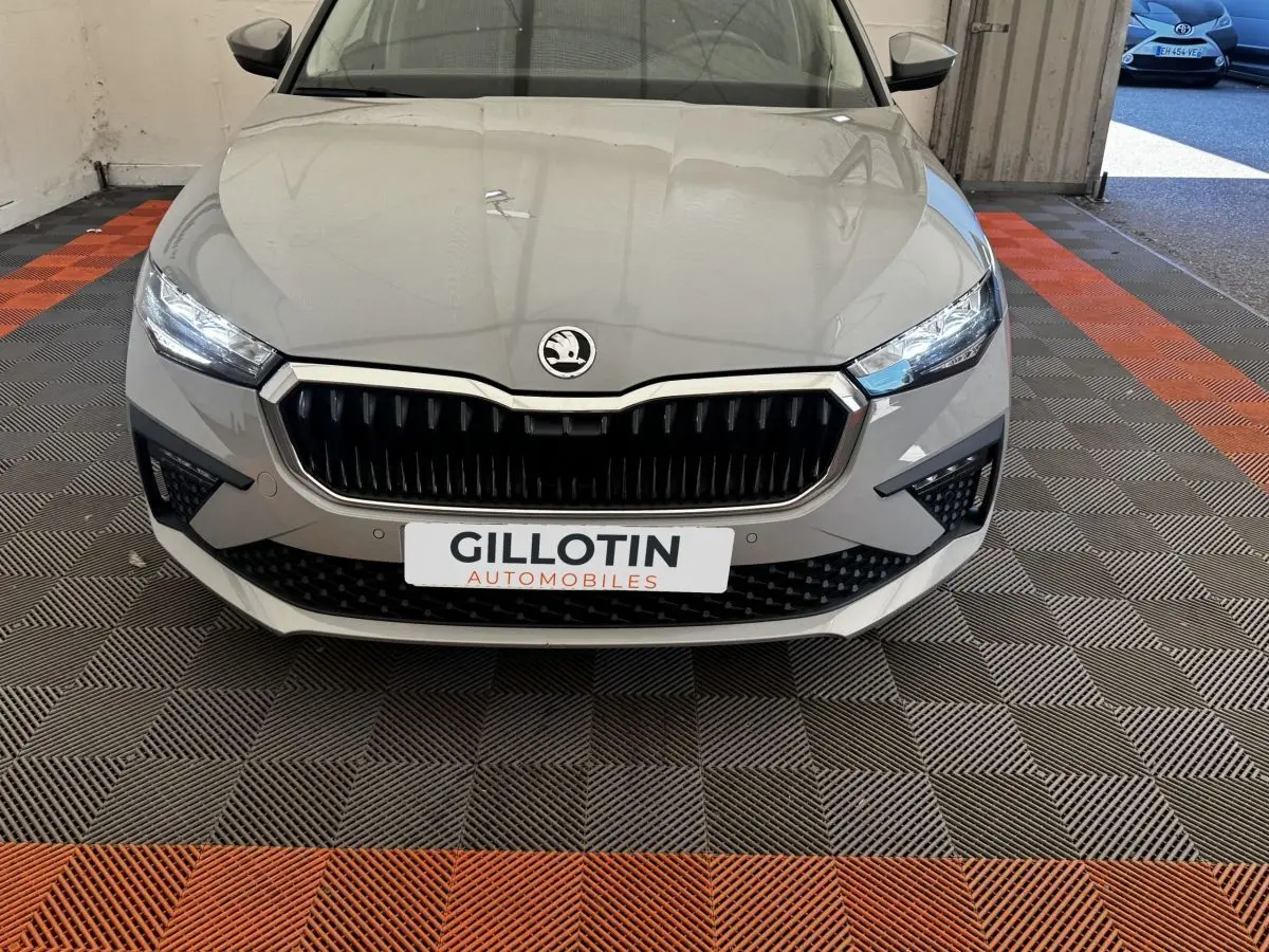 Vue frontale d'une Skoda Scala gris clair avec calandre noire et phares LED allumés sur sol à damier noir et orange.