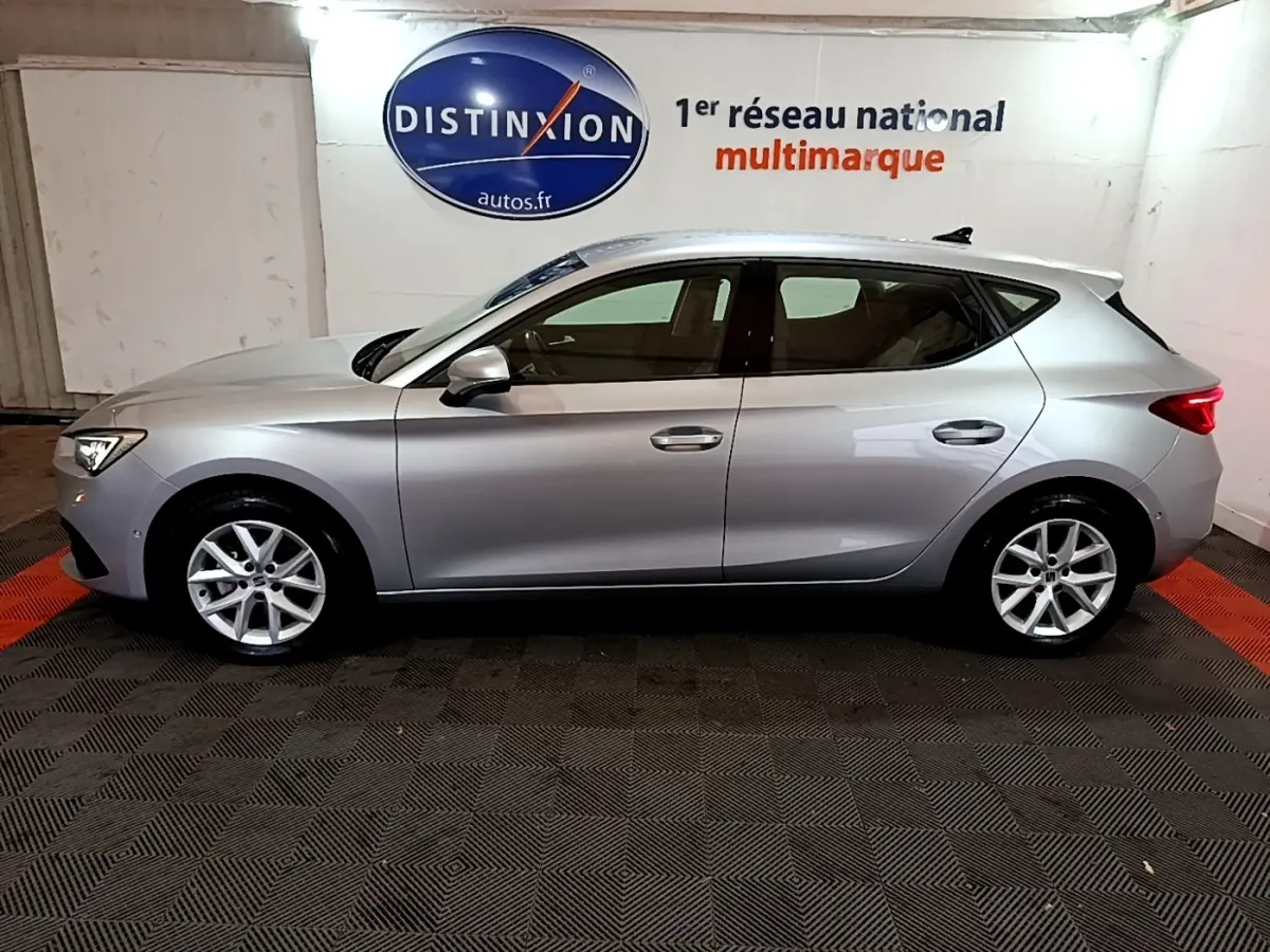 Profil côté gauche d'une SEAT Leon gris clair 2023, en intérieur sur sol damier, avec jantes alliage et feux arrière visibles.