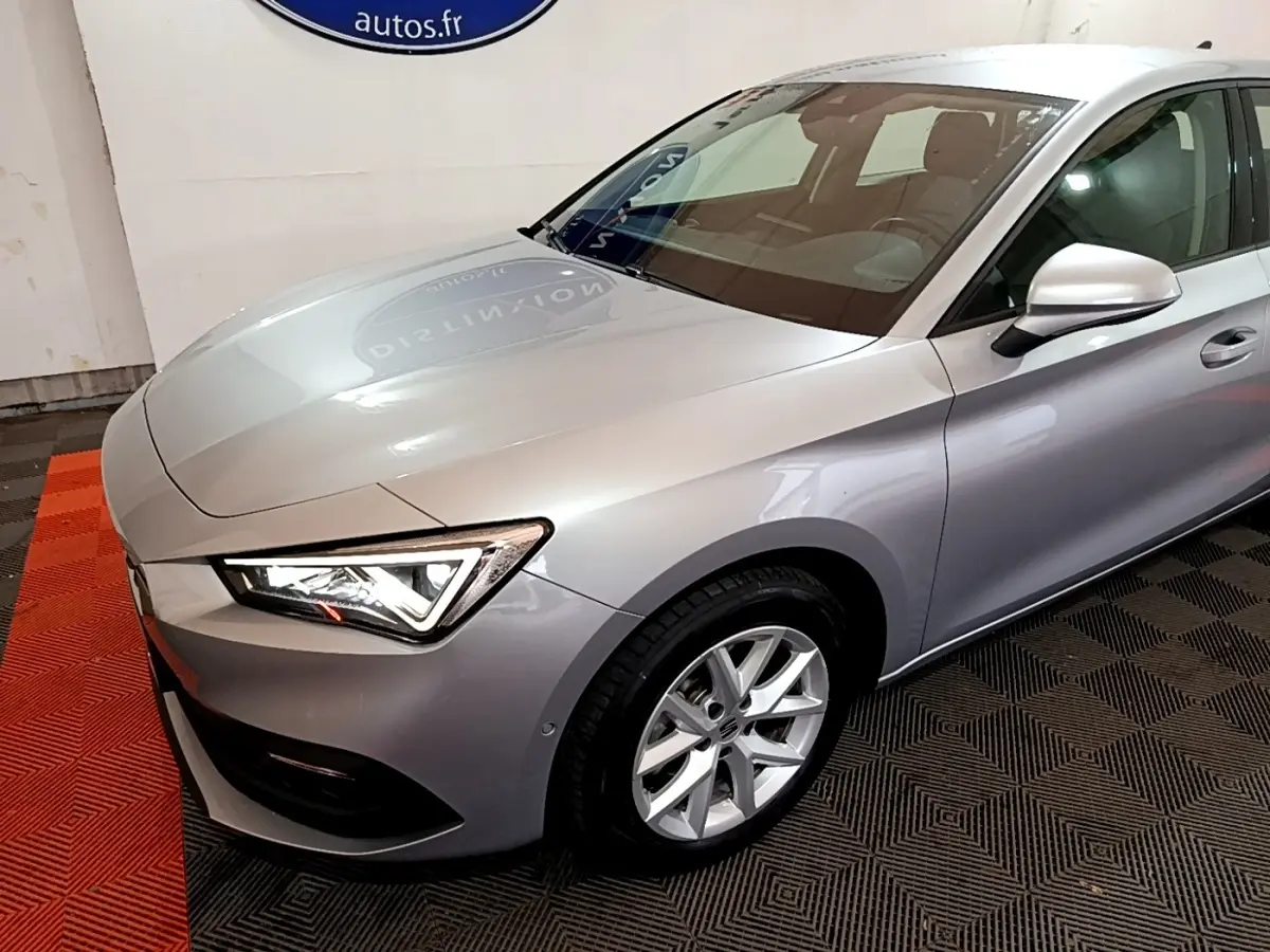 Vue 3/4 avant droit d'une SEAT Leon gris clair avec phares LED et jantes alliage en intérieur showroom.
