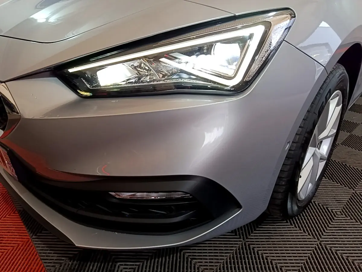 Gros plan sur le phare avant droit allumé d'une SEAT LEON gris foncé 2023 avec jante et pare-chocs noirs.