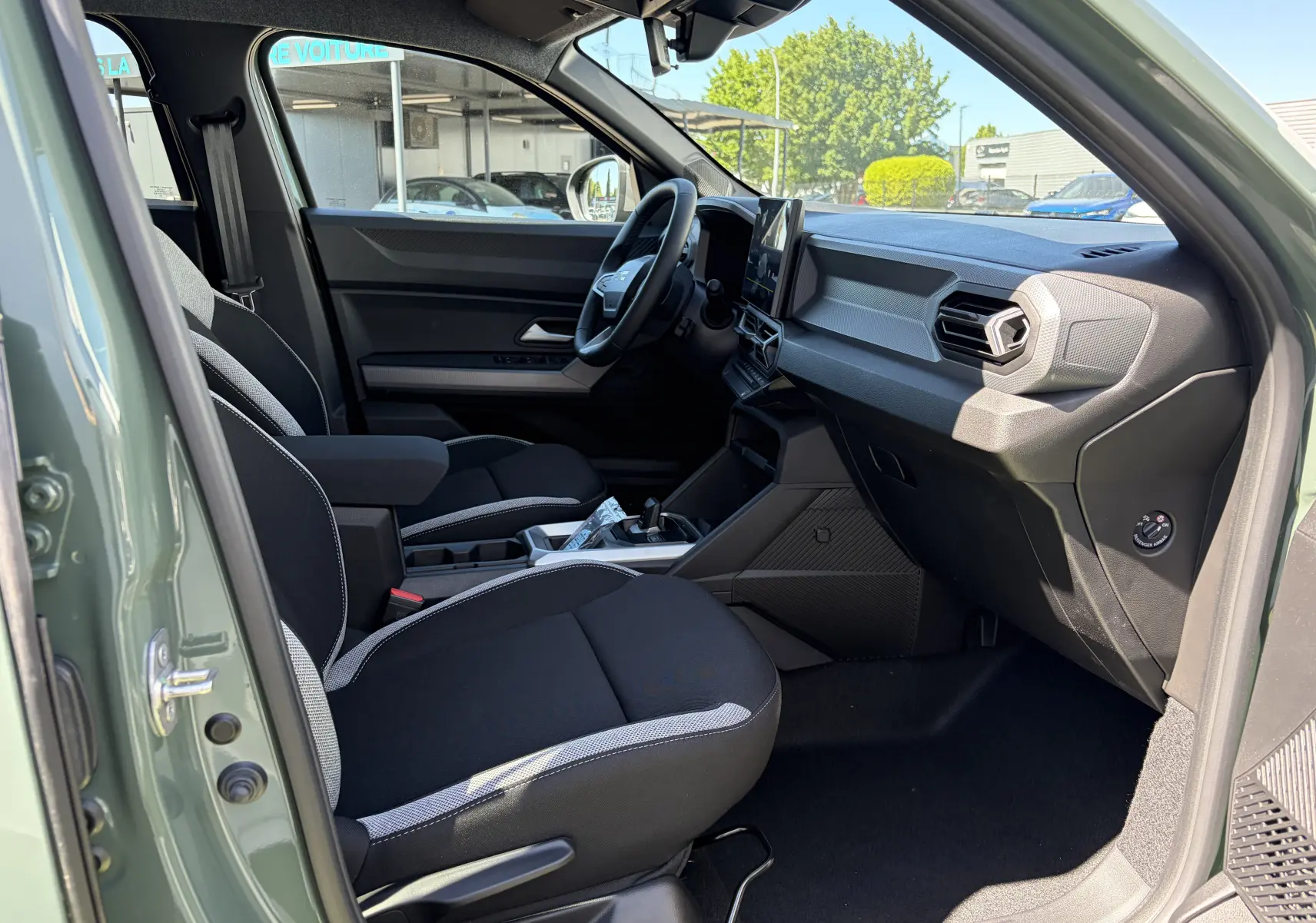 Vue intérieure côté conducteur du Dacia Duster III 1.8 Hybrid 155 kaki lichen, montrant sièges noirs et tableau de bord moderne.