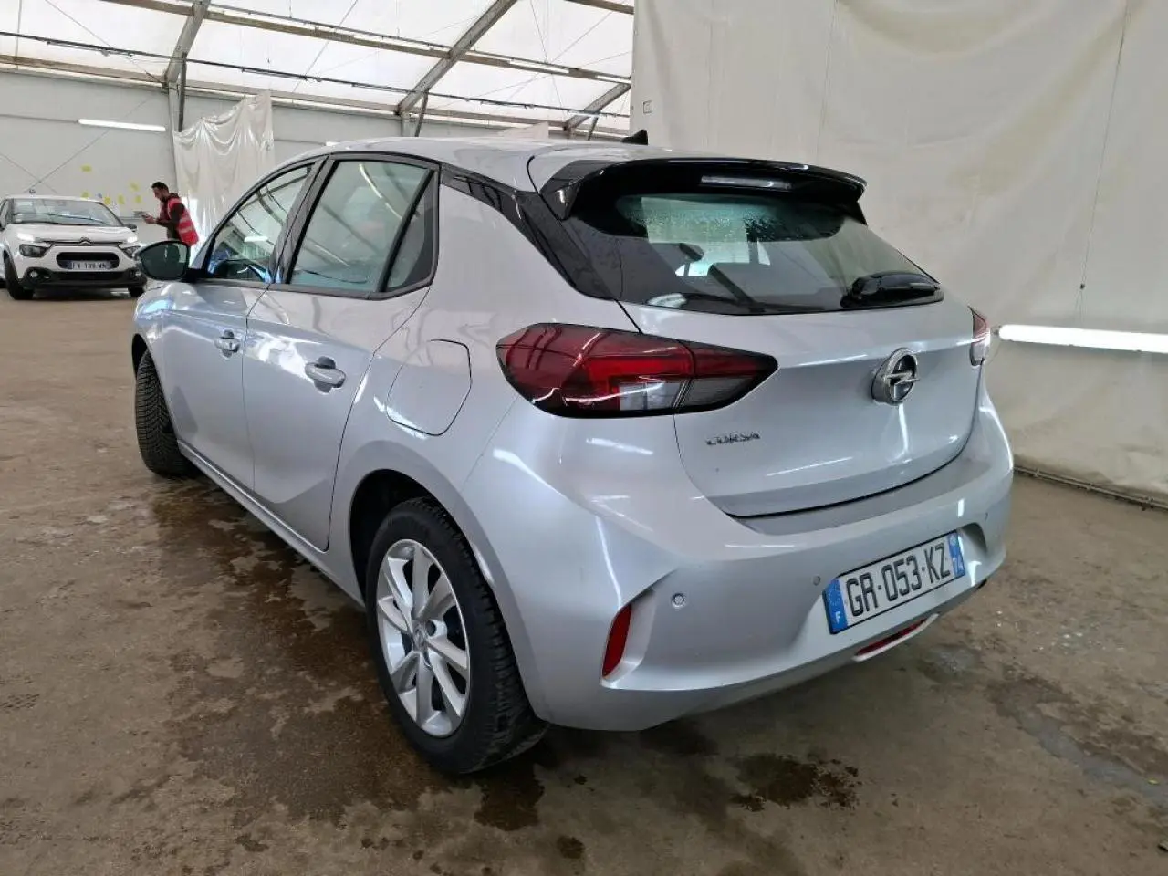 Vue 3/4 arrière gauche d'une Opel Corsa gris clair avec jantes alliage 16'' et rétroviseurs noirs dans un garage.