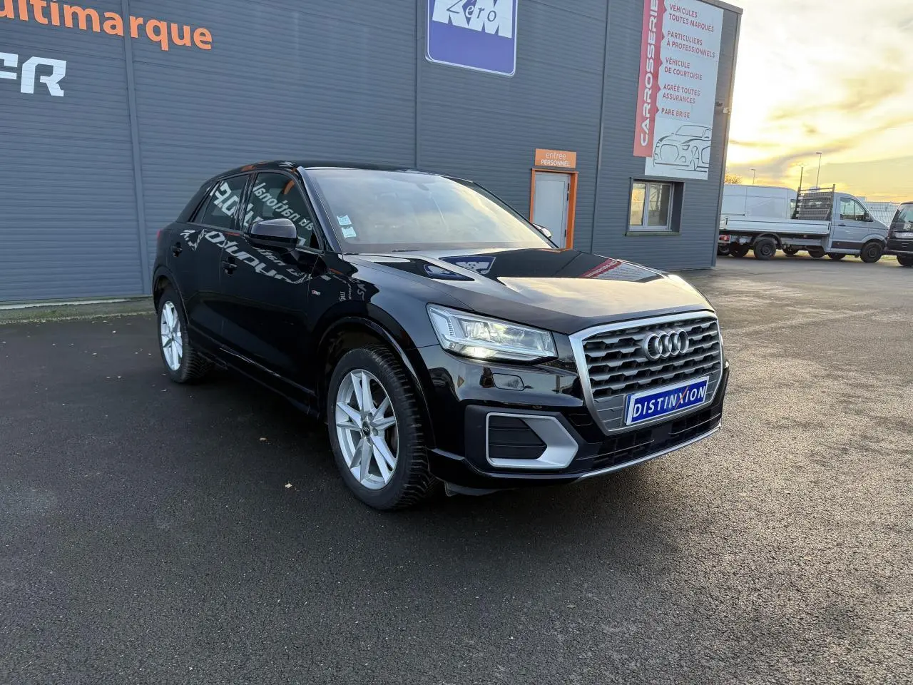 Audi Q2 S Line noir brillant vue 3/4 avant droit sur parking devant bâtiment commercial.