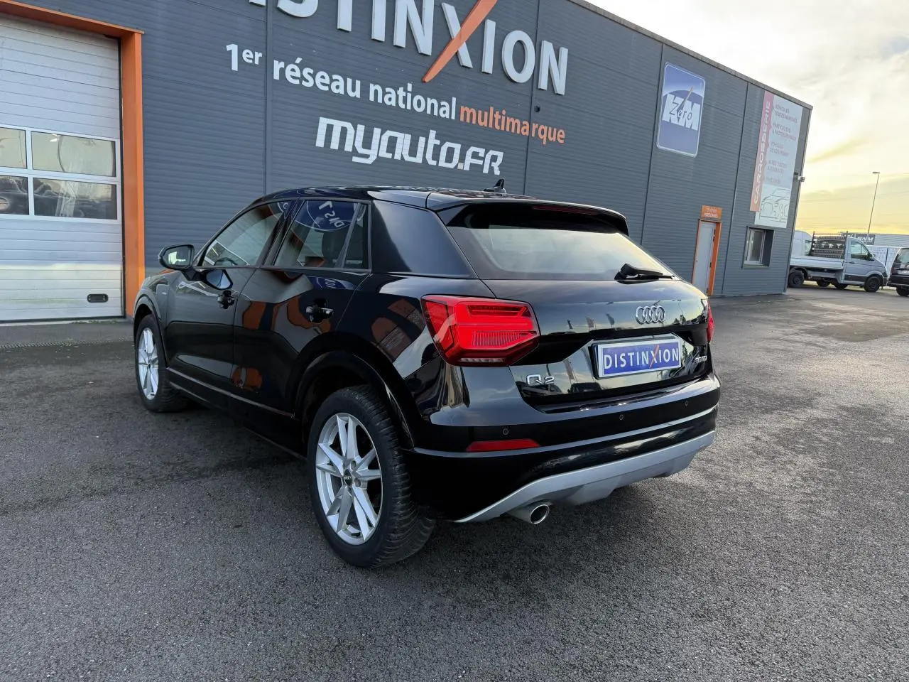 Audi Q2 noir brillant vue 3/4 arrière droit, avec feux rouges et jantes argentées visibles devant un bâtiment commercial.