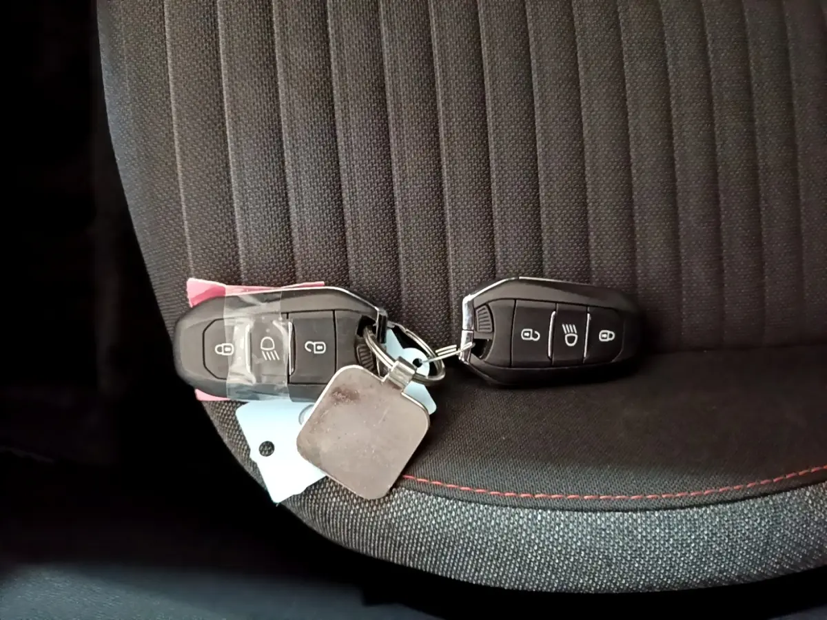 Gros plan sur deux clés noires posées sur un siège tissu gris foncé avec surpiqûres rouges d'une Peugeot 208 électrique blanche.