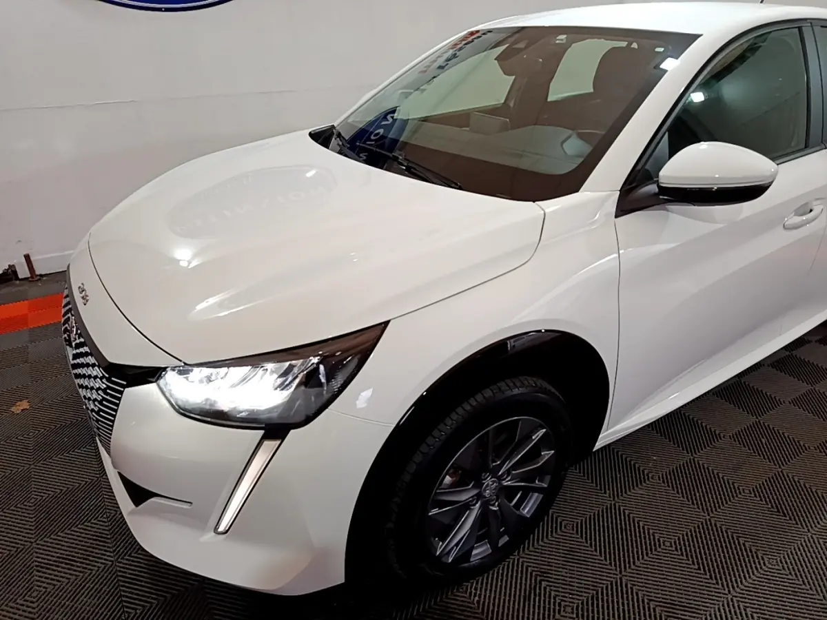 Vue 3/4 avant droit d'une Peugeot 208 électrique Active Business blanche avec phares LED allumés et jantes foncées.