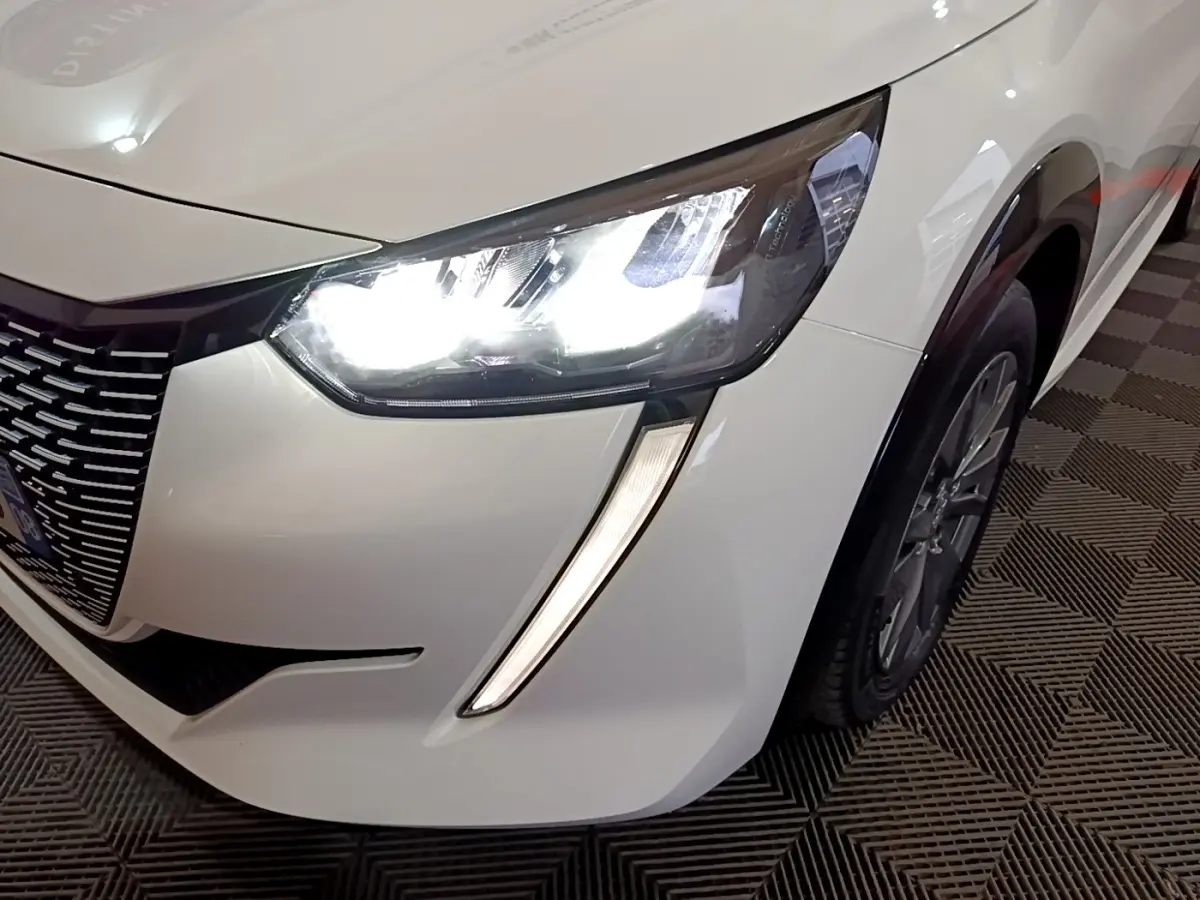 Gros plan sur l'avant droit blanc d'une Peugeot 208 électrique avec phare LED allumé et jante visible.