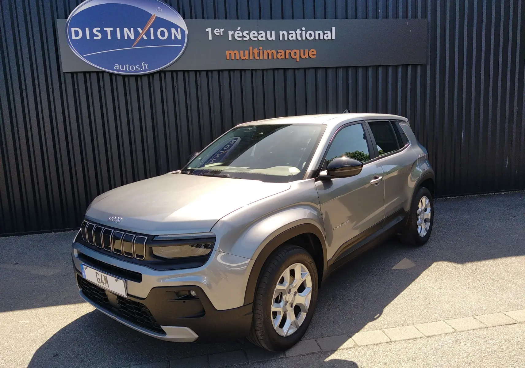 Jeep Avenger eHYBRID gris en 3/4 avant droit, avec calandre distinctive et jantes argentées, stationné devant un mur noir.