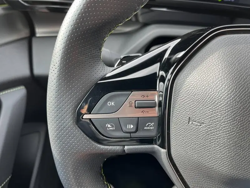 Gros plan sur les commandes multifonctions du volant côté gauche d'une Peugeot 308 SW 2023 en cuir perforé noir avec surpiqûres vertes.