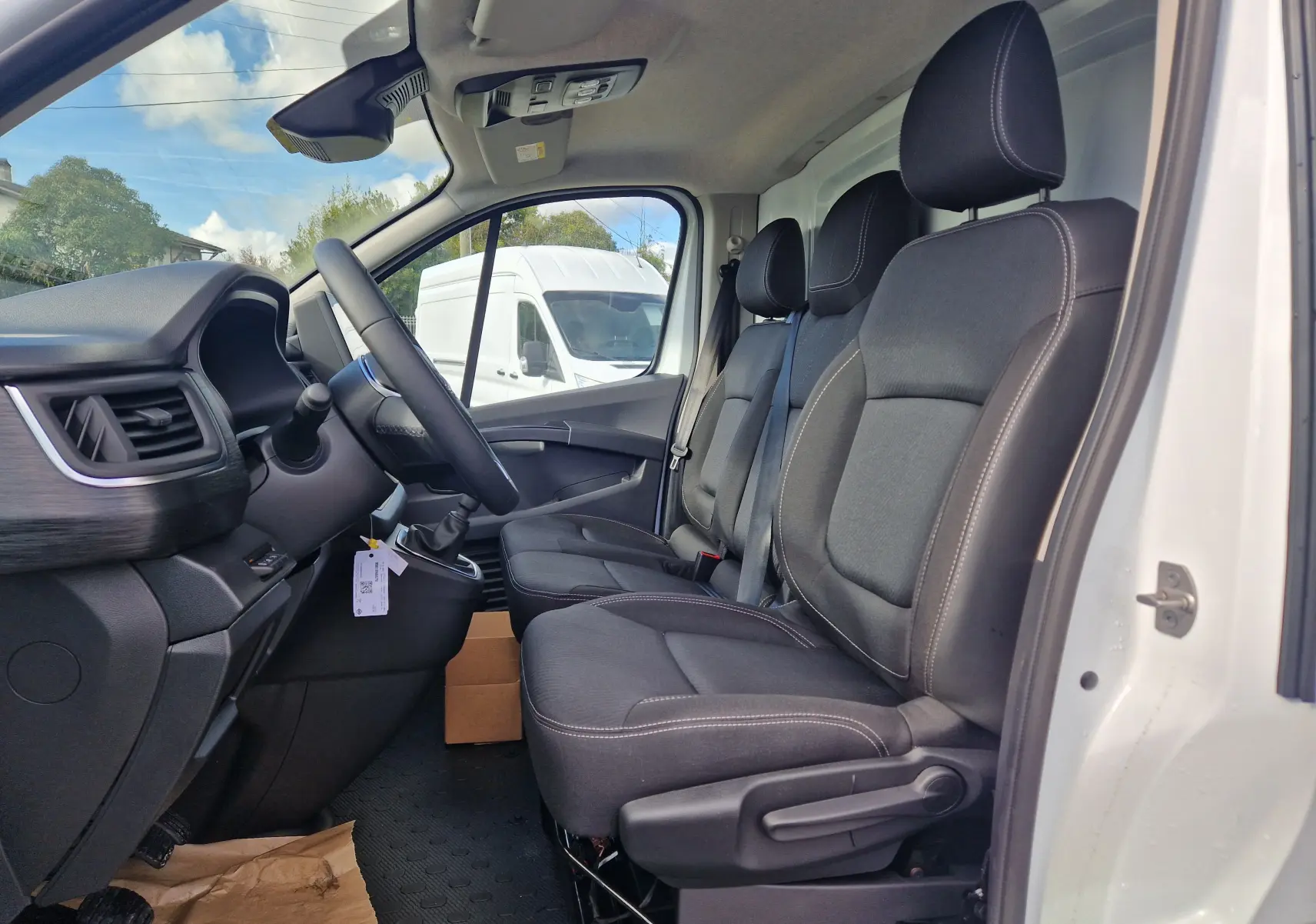 Vue intérieure côté conducteur du Nissan Primastar blanc 2025, montrant les trois sièges avant en tissu noir et le tableau de bord.