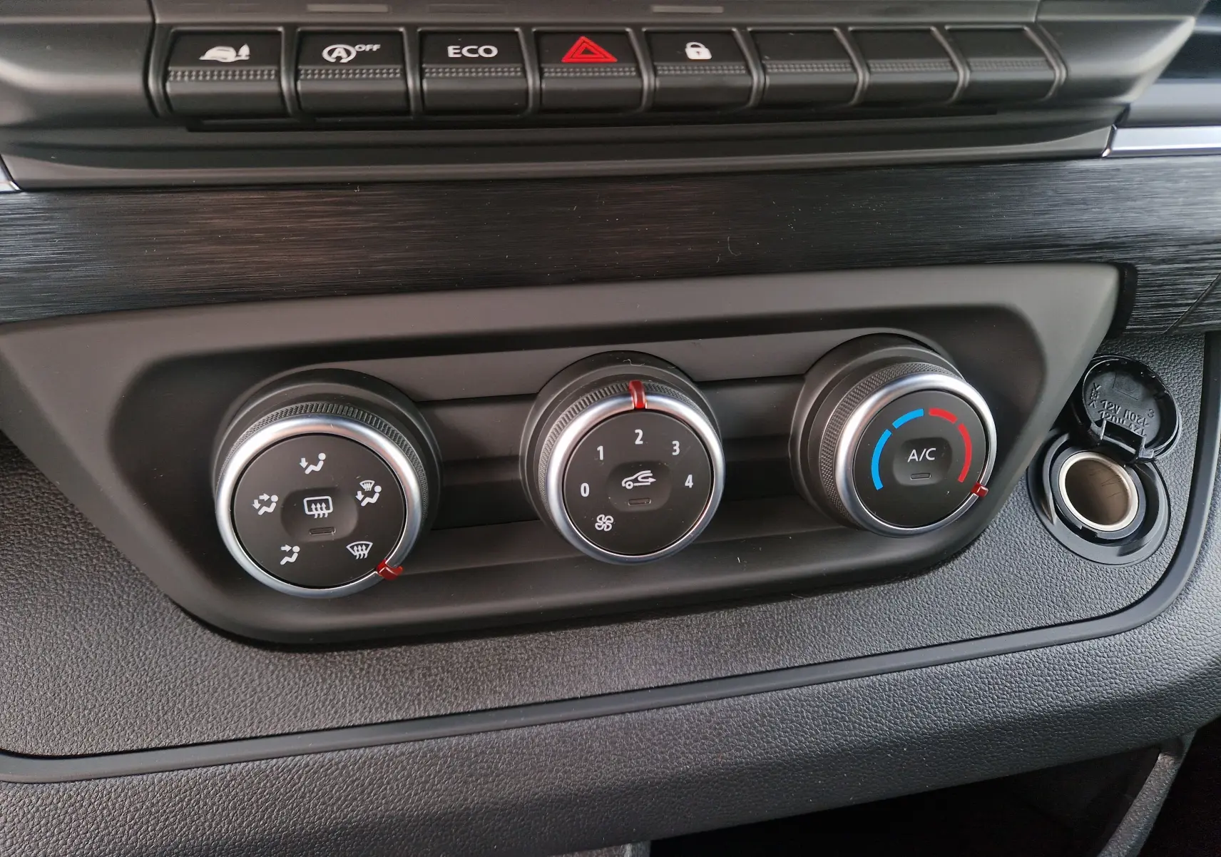 Gros plan sur les commandes de climatisation noires du Nissan Primastar blanc, avec boutons rotatifs et prise 12V ouverte.