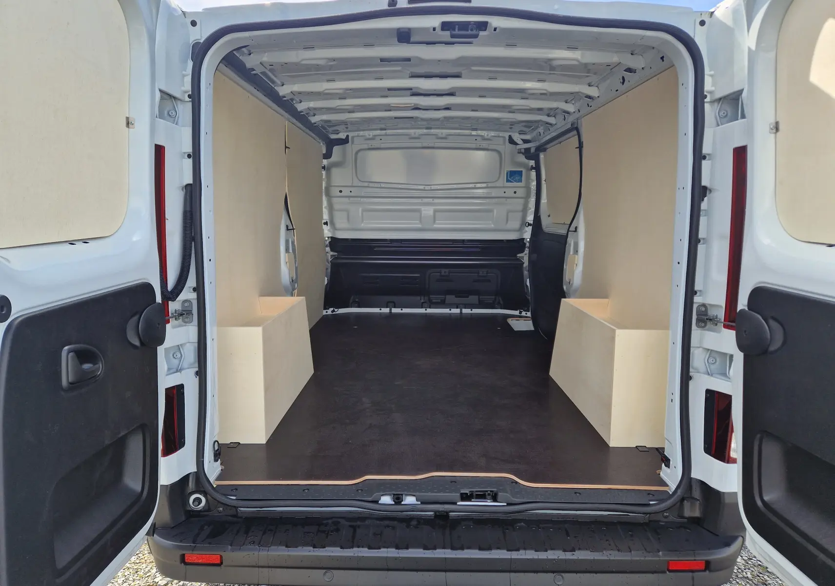 Vue arrière ouverte du Nissan Primastar blanc 2025, montrant l'intérieur cargo aménagé avec panneaux bois et sol noir antidérapant.