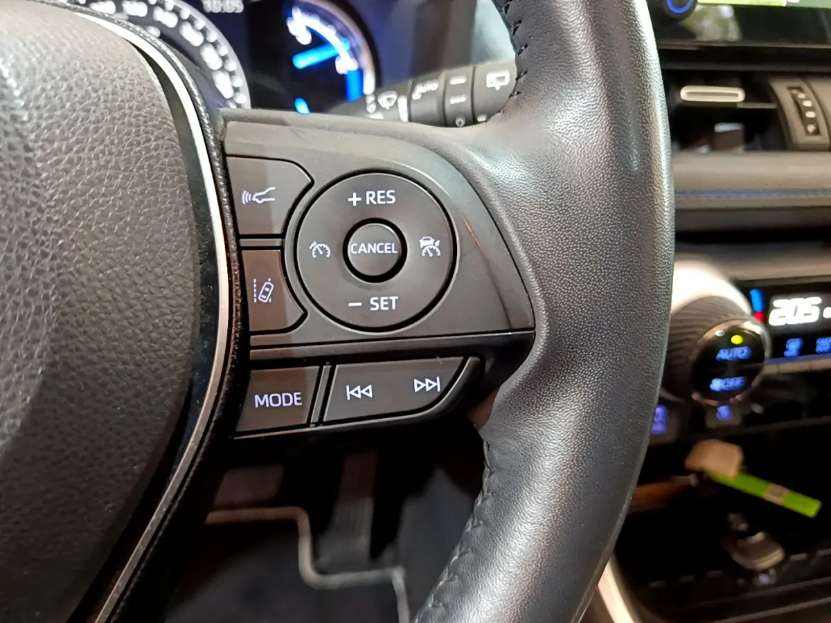 Gros plan sur les commandes du volant en cuir noir du Toyota RAV4 2WD 218ch Collection, avec tableau de bord lumineux en arrière-plan.
