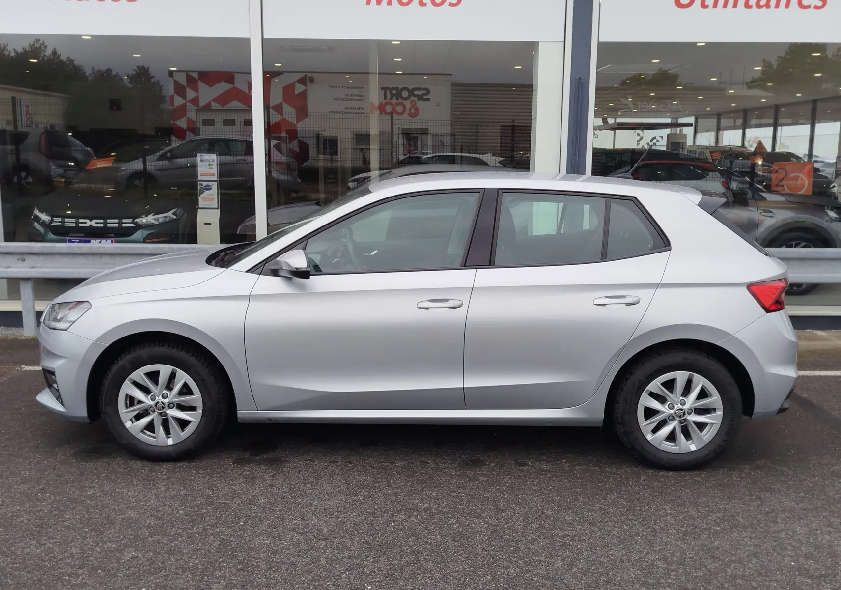 Vue de profil côté gauche d'une Skoda Fabia gris argent métallisé 2023 garée devant une vitrine de concession.