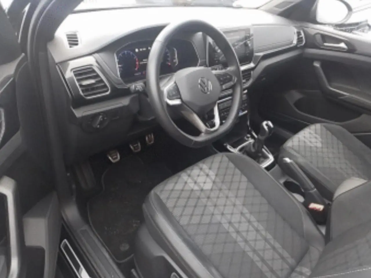 Vue intérieure côté conducteur du Volkswagen T-Cross 2025 noir métal, avec volant multifonction, sièges tissu à motif losanges et levier de vitesse manuel.