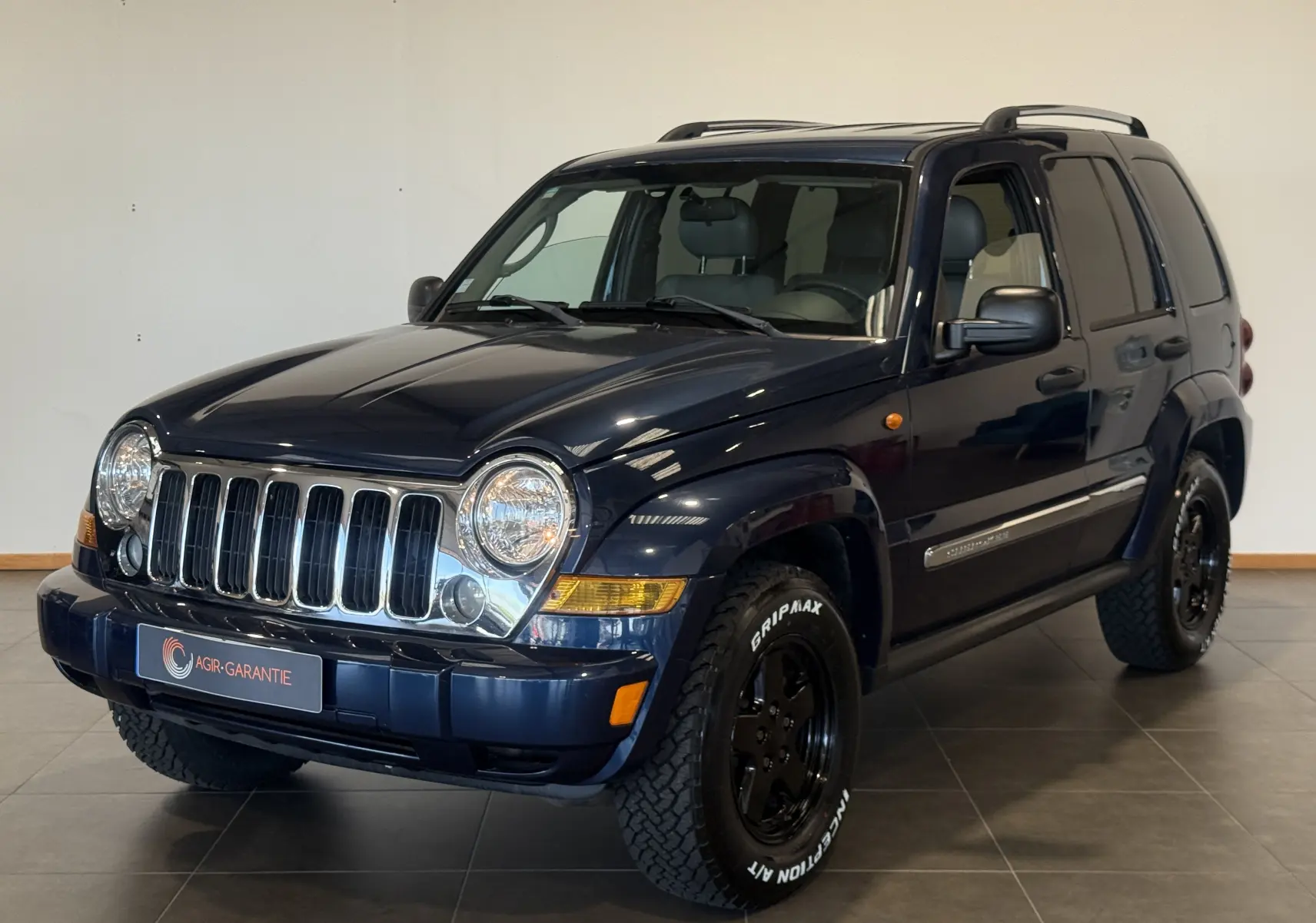 Jeep Cherokee bleu 2005 en 3/4 avant droit, avec calandre chromée et pneus tout-terrain marqués Gripmax.