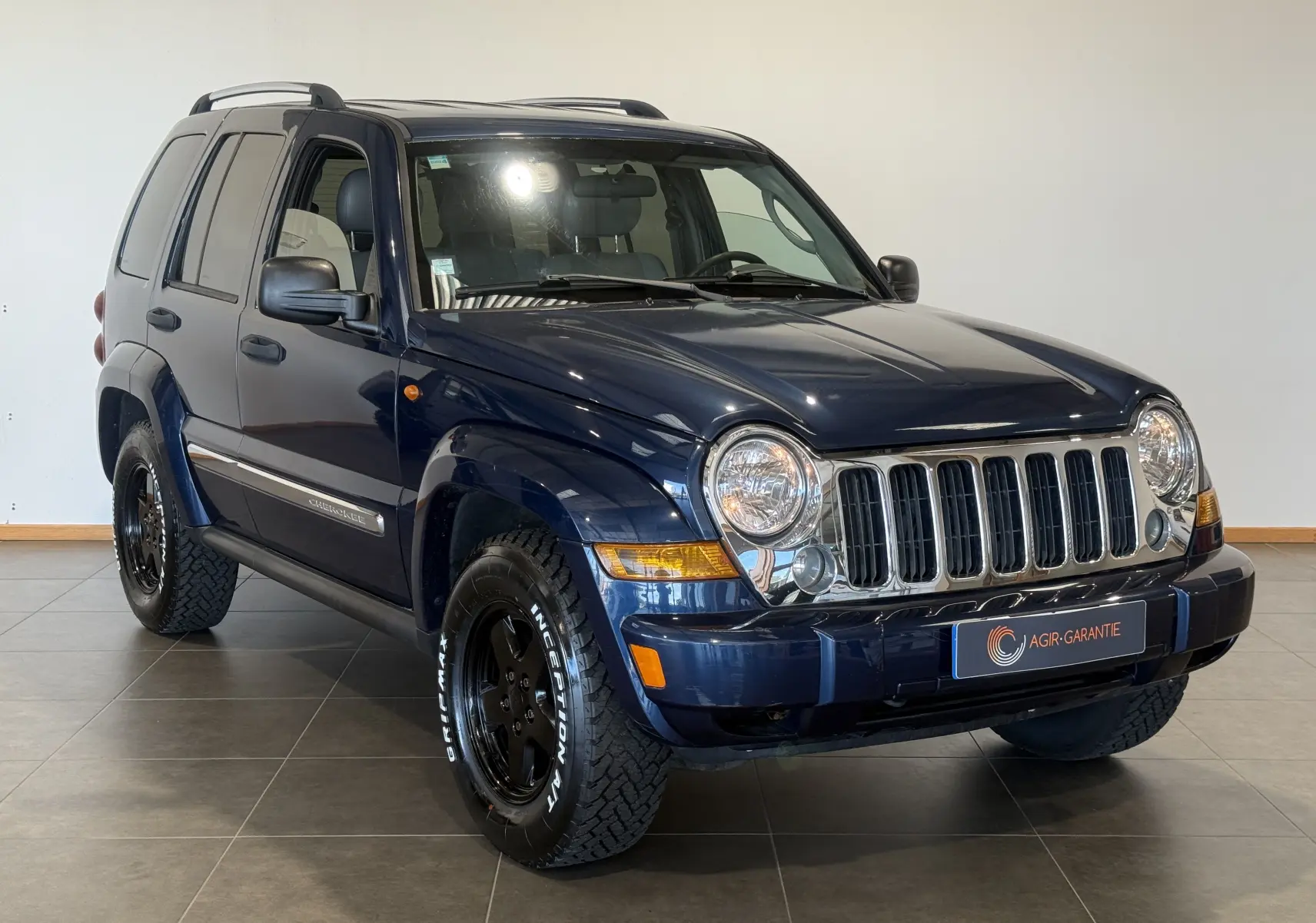 Jeep Cherokee bleu 2005 en 3/4 avant droit, avec calandre chromée et jantes noires tout-terrain.