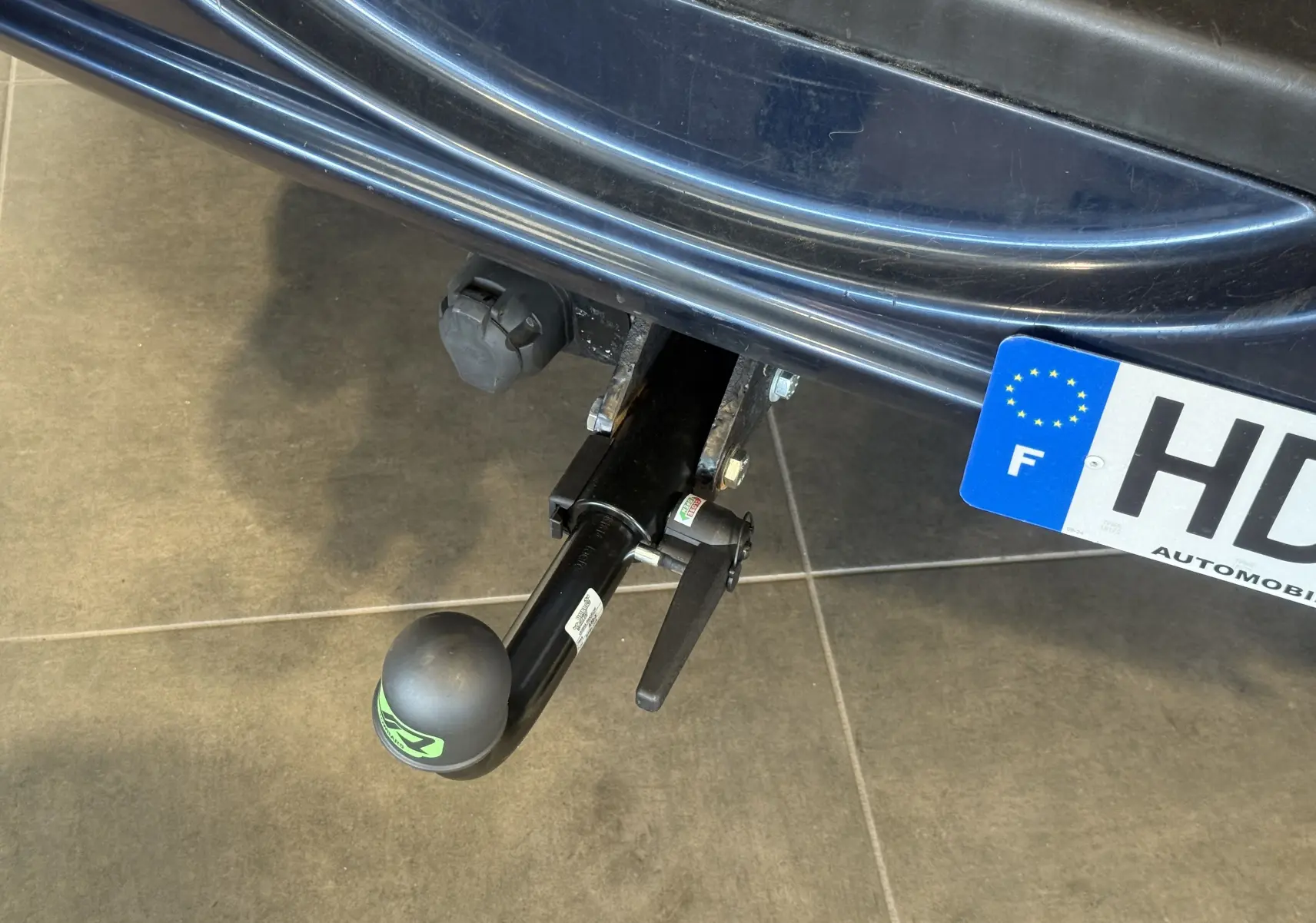 Gros plan sur l'attelage arrière noir d'un Jeep Cherokee bleu 2005, avec une prise électrique visible à côté du pare-chocs.