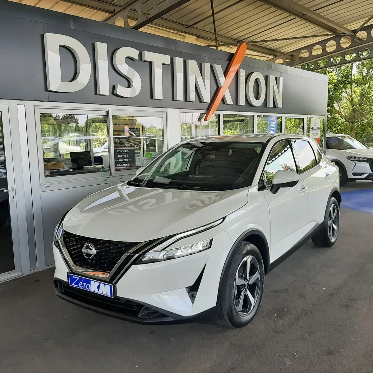 Nissan Qashqai blanc lunaire en 3/4 avant droit, avec calandre noire et jantes alliage distinctives, sous un auvent showroom.