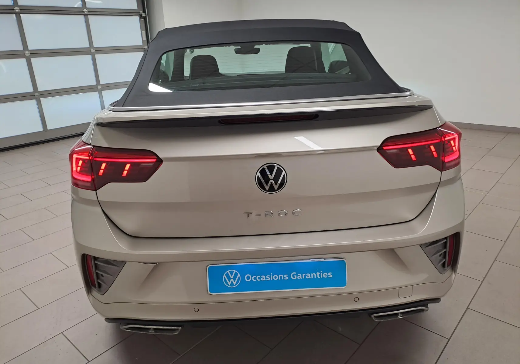 Vue arrière du Volkswagen T-Roc Cabriolet gris ivoire avec capote noire et feux arrière allumés dans un showroom.