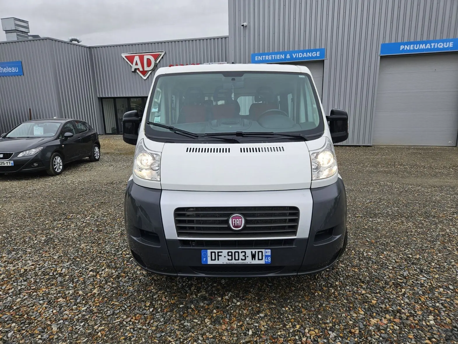 Vue de face d'un Fiat Ducato Combi blanc avec pare-chocs noir, immatriculé en France, stationné devant un garage.