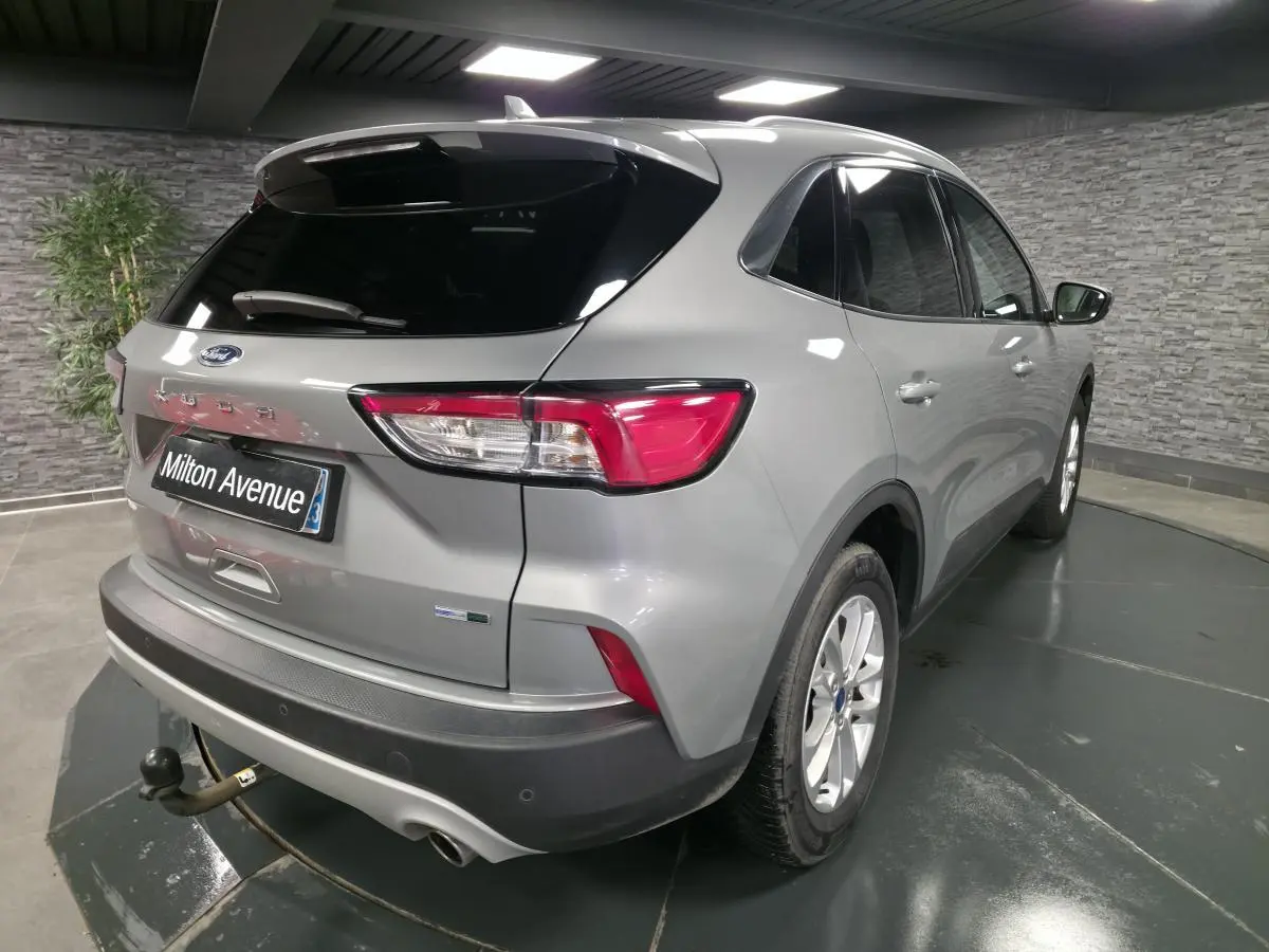 Vue 3/4 arrière droit d'un Ford Kuga gris métallisé avec crochet d'attelage et vitres arrière surteintées en intérieur showroom.