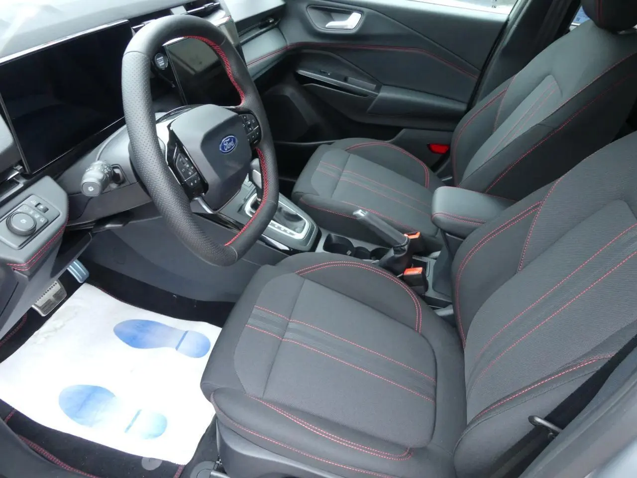 Intérieur avant du Ford Puma 2025, sièges en tissu gris avec surpiqûres rouges et volant sport multifonction.