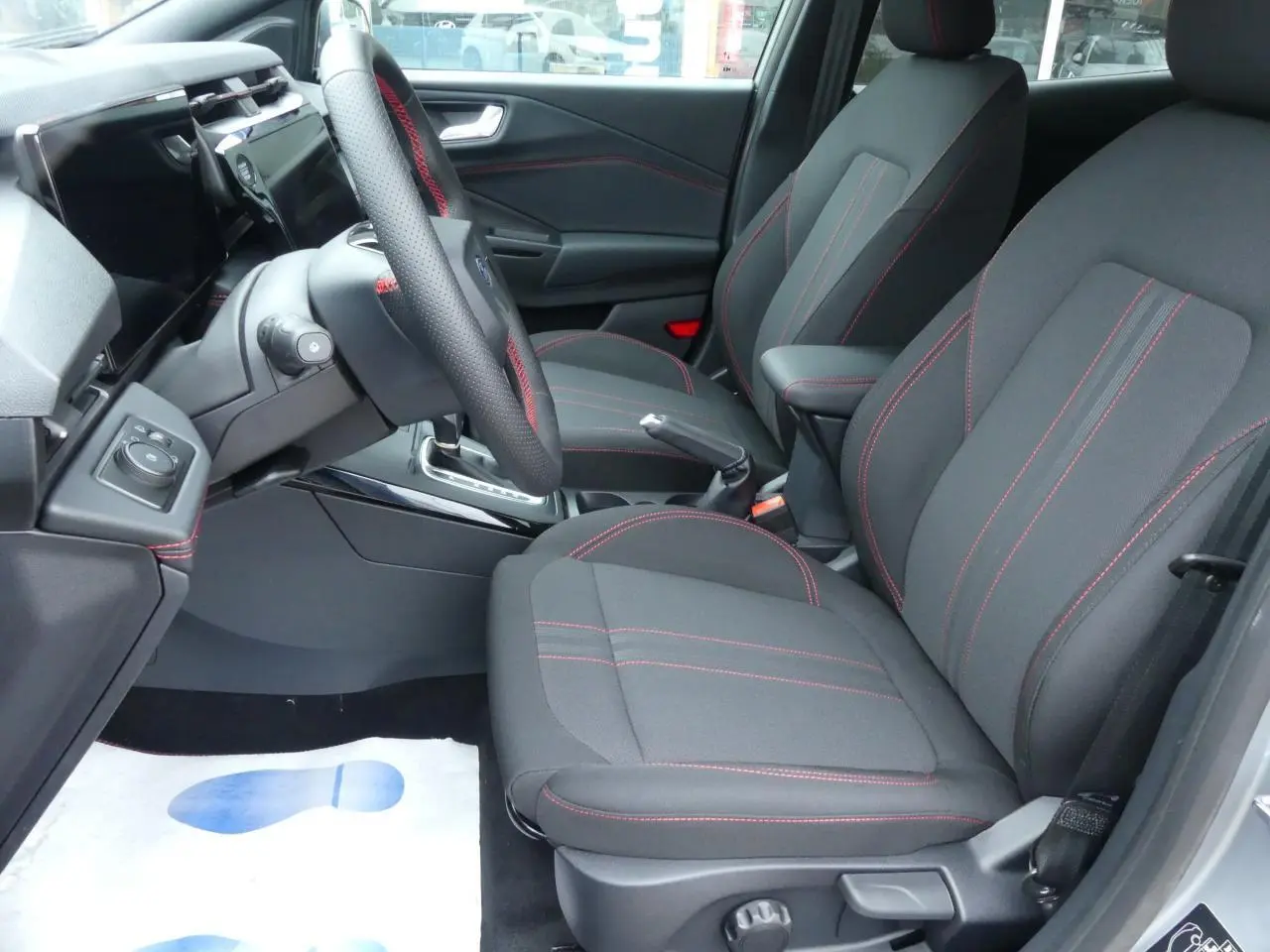 Vue intérieure côté conducteur du Ford Puma 2025, sièges tissu noirs avec surpiqûres rouges et volant sport.