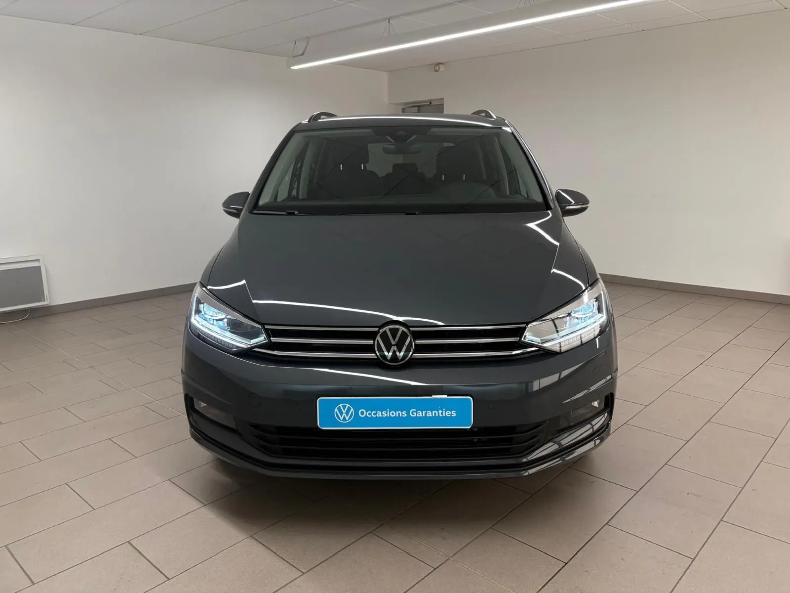 Volkswagen Touran gris dauphin vu de face en intérieur, avec phares LED allumés et calandre chromée.