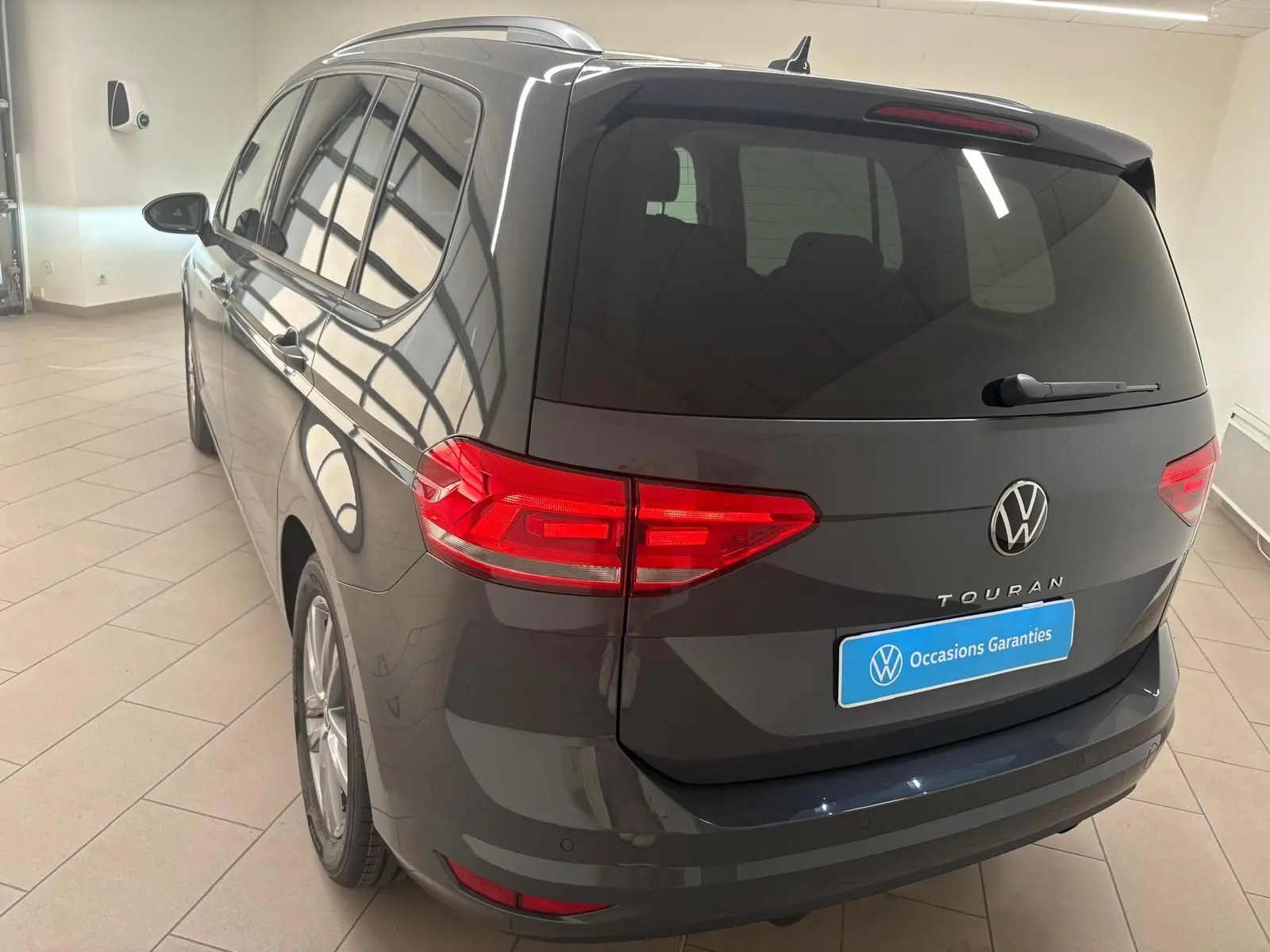 Vue 3/4 arrière droite d'un Volkswagen Touran gris dauphin 2025 avec feux arrière LED et hayon fermé dans un showroom.