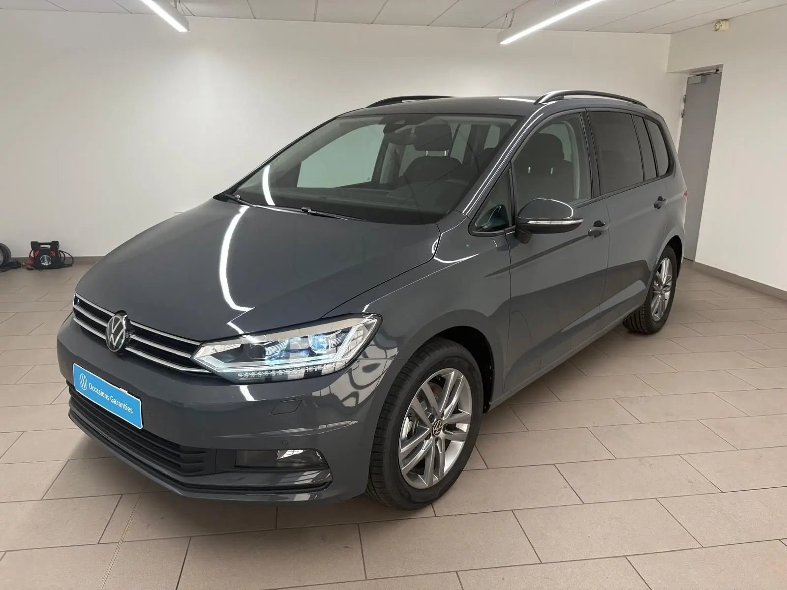 Volkswagen Touran gris dauphin en 3/4 avant droit dans un intérieur, avec jantes alliage et phares LED allumés.