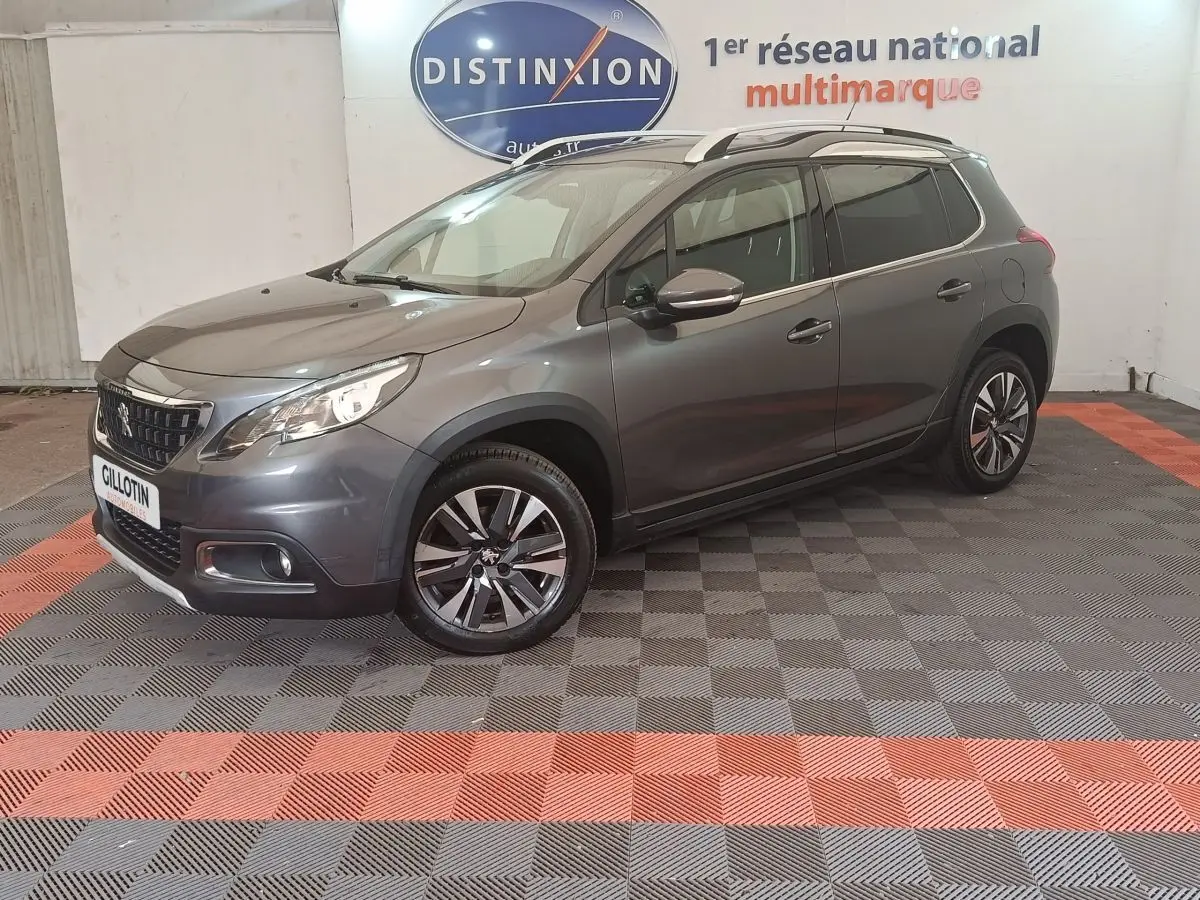 Peugeot 2008 gris foncé en 3/4 avant droit, avec jantes alliage et barres de toit chromées visibles.