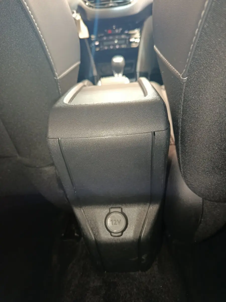 Vue intérieure arrière centrée sur la console centrale noire avec prise 12V entre les sièges avant du Peugeot 2008 gris foncé.