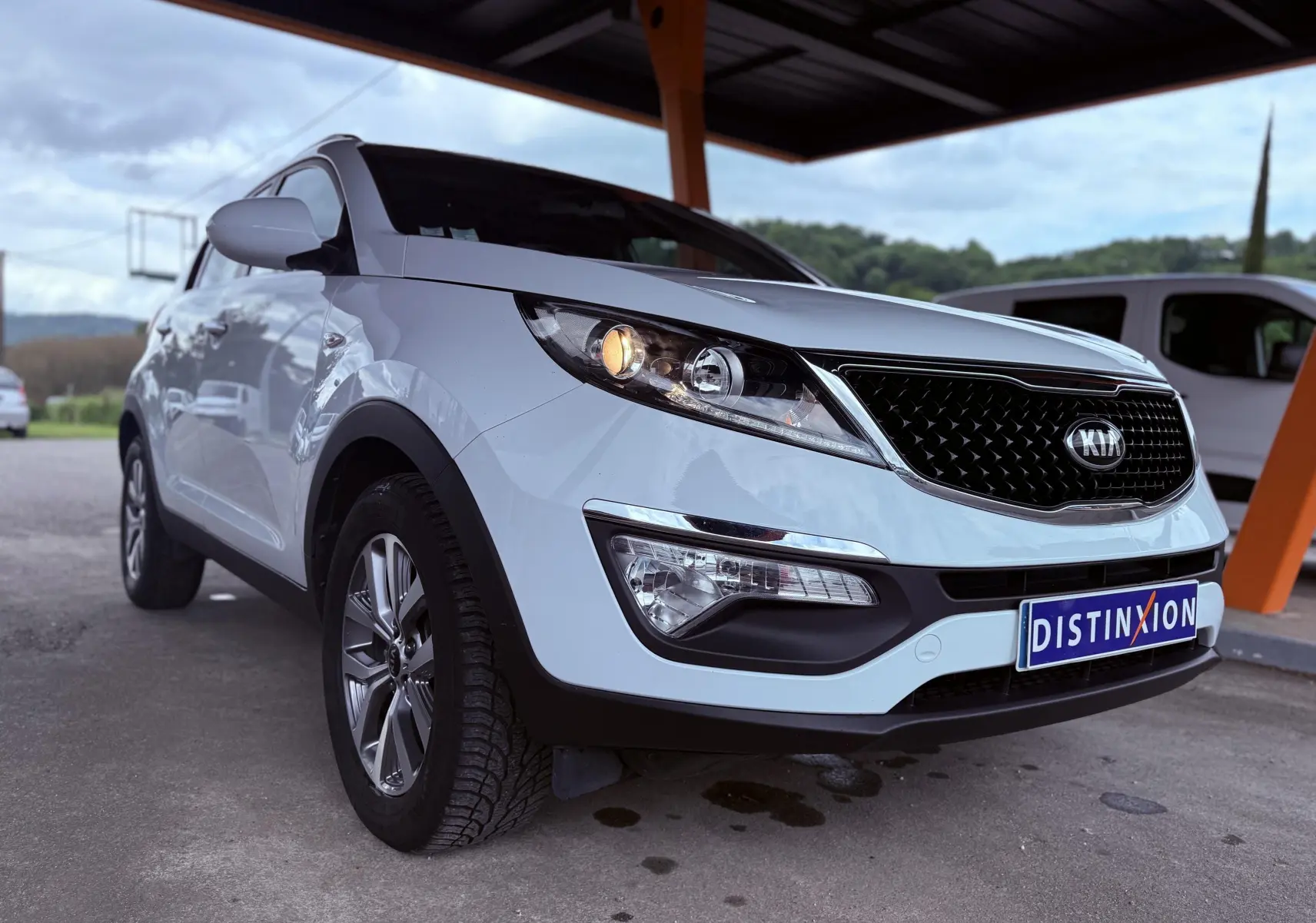 KIA Sportage blanche vue en 3/4 avant droit sous un abri avec phare avant allumé et plaque Distinxion.