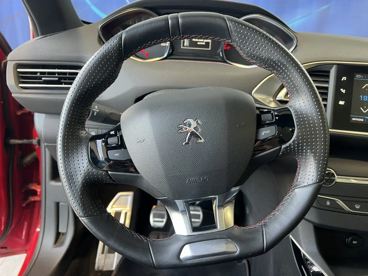 Vue rapprochée du volant cuir perforé avec coutures rouges de la Peugeot 308 GT Line rouge, tableau de bord en arrière-plan.
