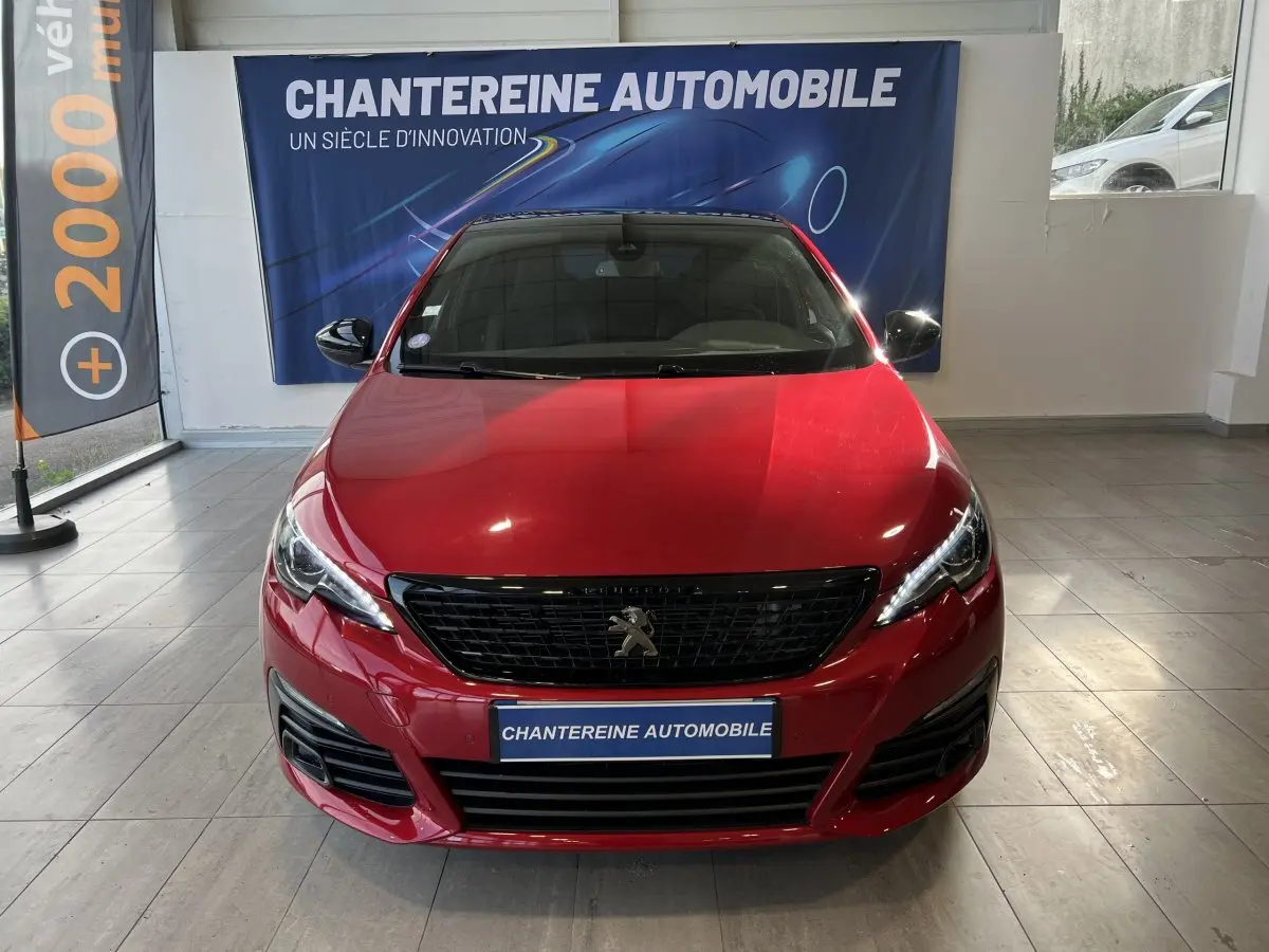 Vue frontale d'une Peugeot 308 Puretech 130 S&S GT Line rouge brillante en showroom avec calandre noire et phares LED.